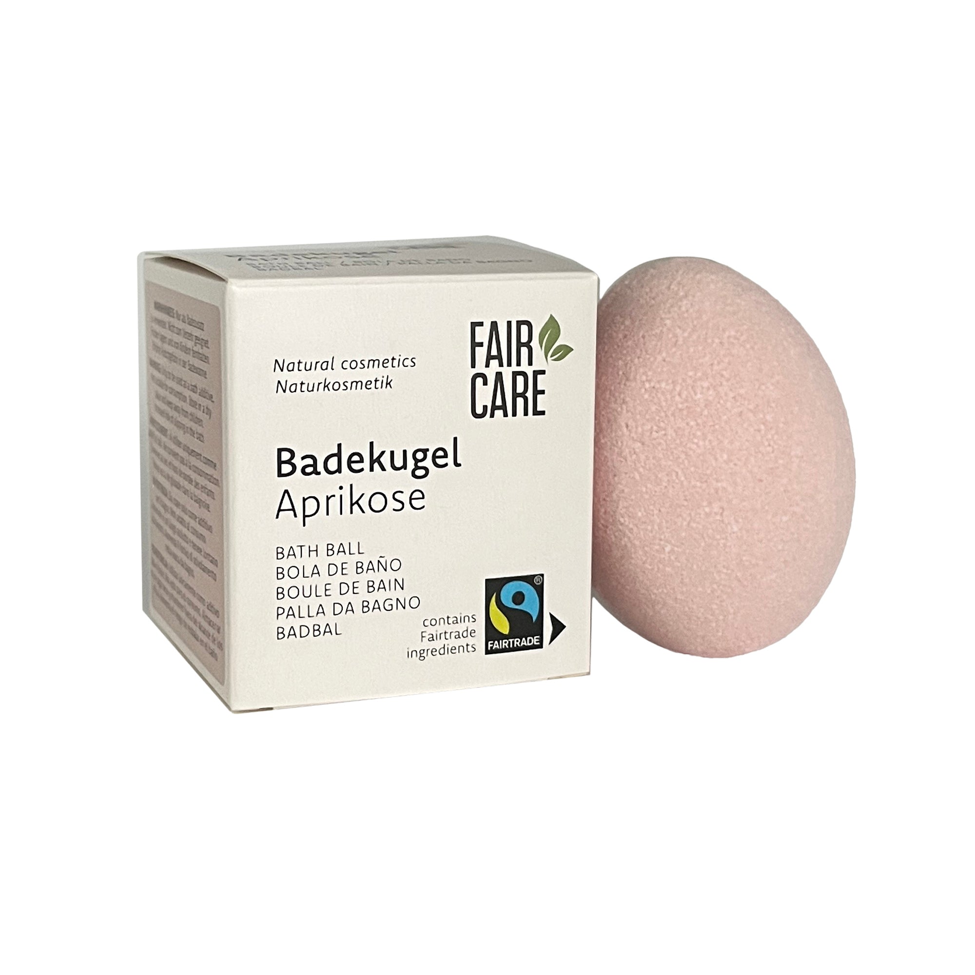 Eine blassrosa Badekugel liegt neben einer weißen Schachtel mit der Aufschrift Fair Care Badekugel Aprikose mit Fairtrade- und Naturkosmetiksymbolen und mehrsprachigen Beschreibungen.