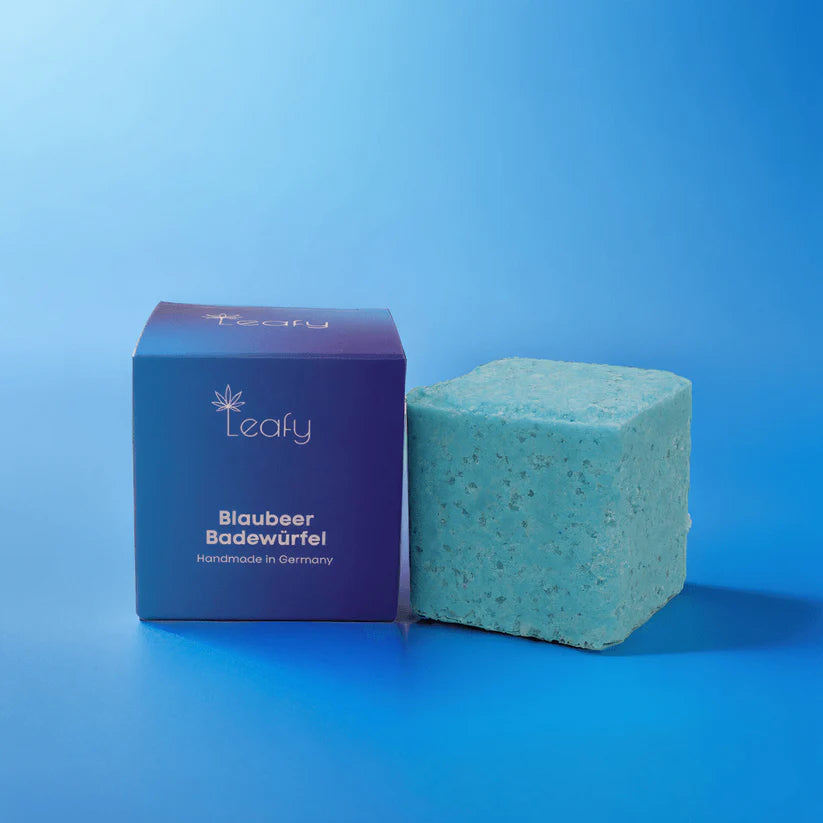 Eine blaue quadratische Badebombe sitzt neben der passenden lila-blauen Schachtel mit der Aufschrift "Leafy Blaubeer Badewürfel Handmade in Germany" vor einem blauen Hintergrund mit Farbverlauf.