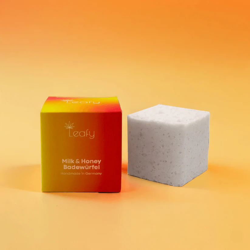 Eine quadratische Schachtel mit der Aufschrift Leafy Milk & Honey Badewürfel steht neben einem weißen Badewürfel mit Flecken vor einem orangefarbenen Hintergrund mit Farbverlauf.