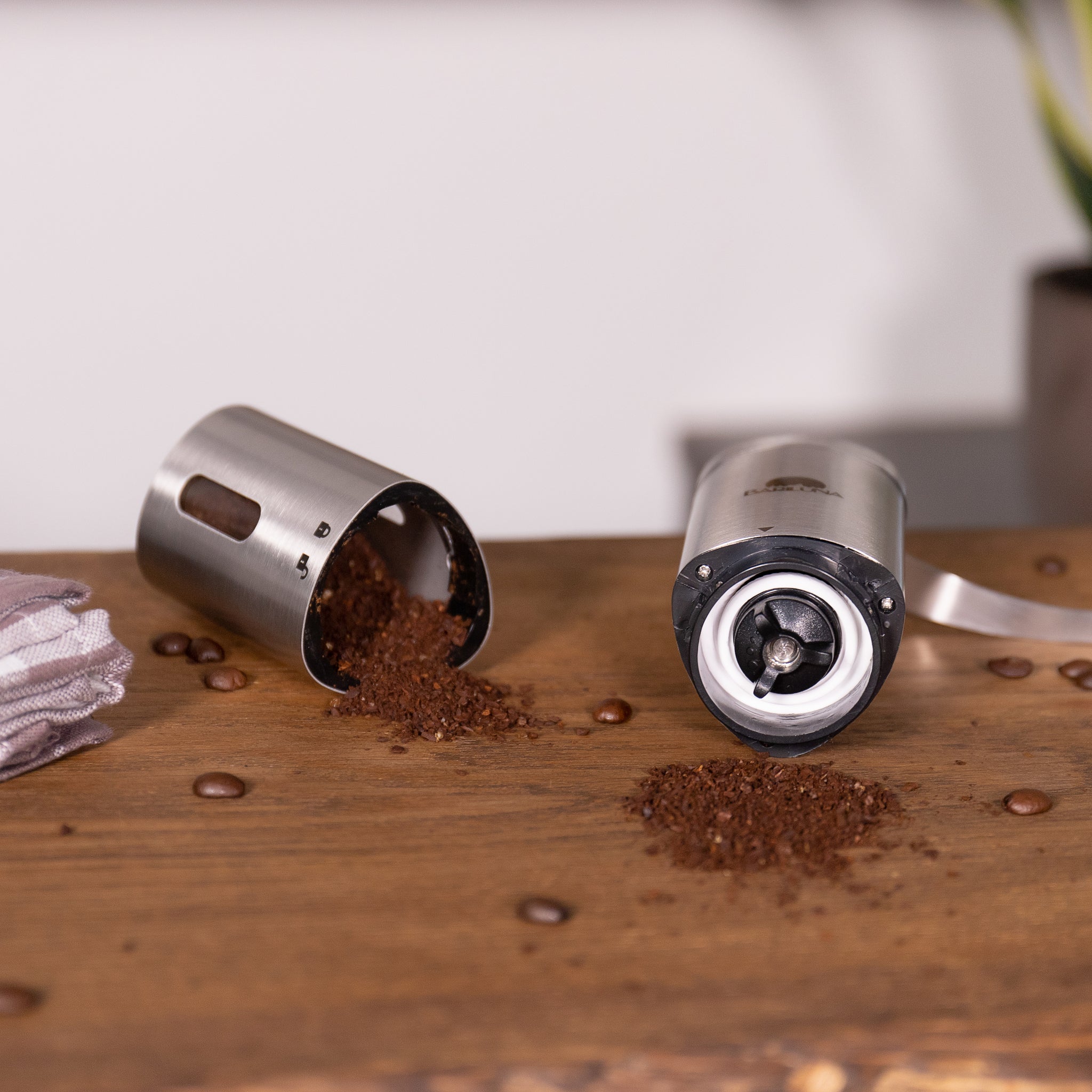 Zerlegbare Kaffeemühle auf Holztisch, mit gemahlenem Kaffee und Kaffeebohnen verstreut; nahe liegendes Tuch.