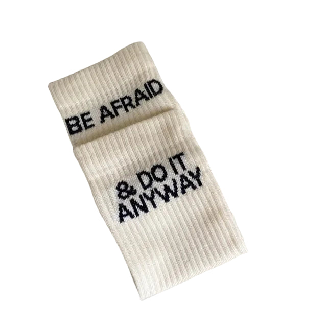 Ein Paar cremefarbene, gerippte Socken mit dem schwarzen Text "BE AFRAID & DO IT ANYWAY" auf einer hellgrauen Oberfläche.
