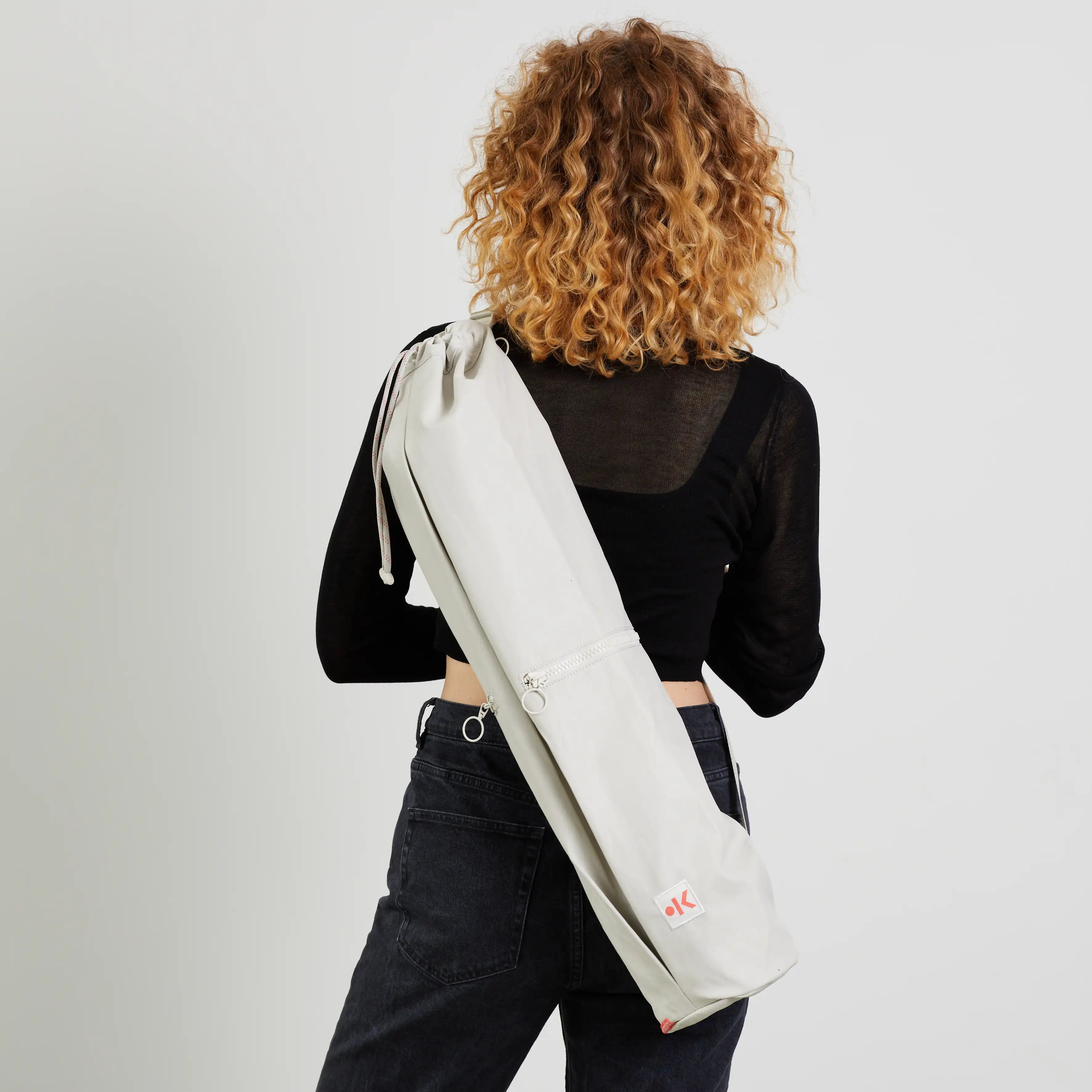 Person mit lockigem Haar trägt eine weiße Tasche über der Schulter, in einem schwarzen Oberteil und Jeans vor weißem Hintergrund.