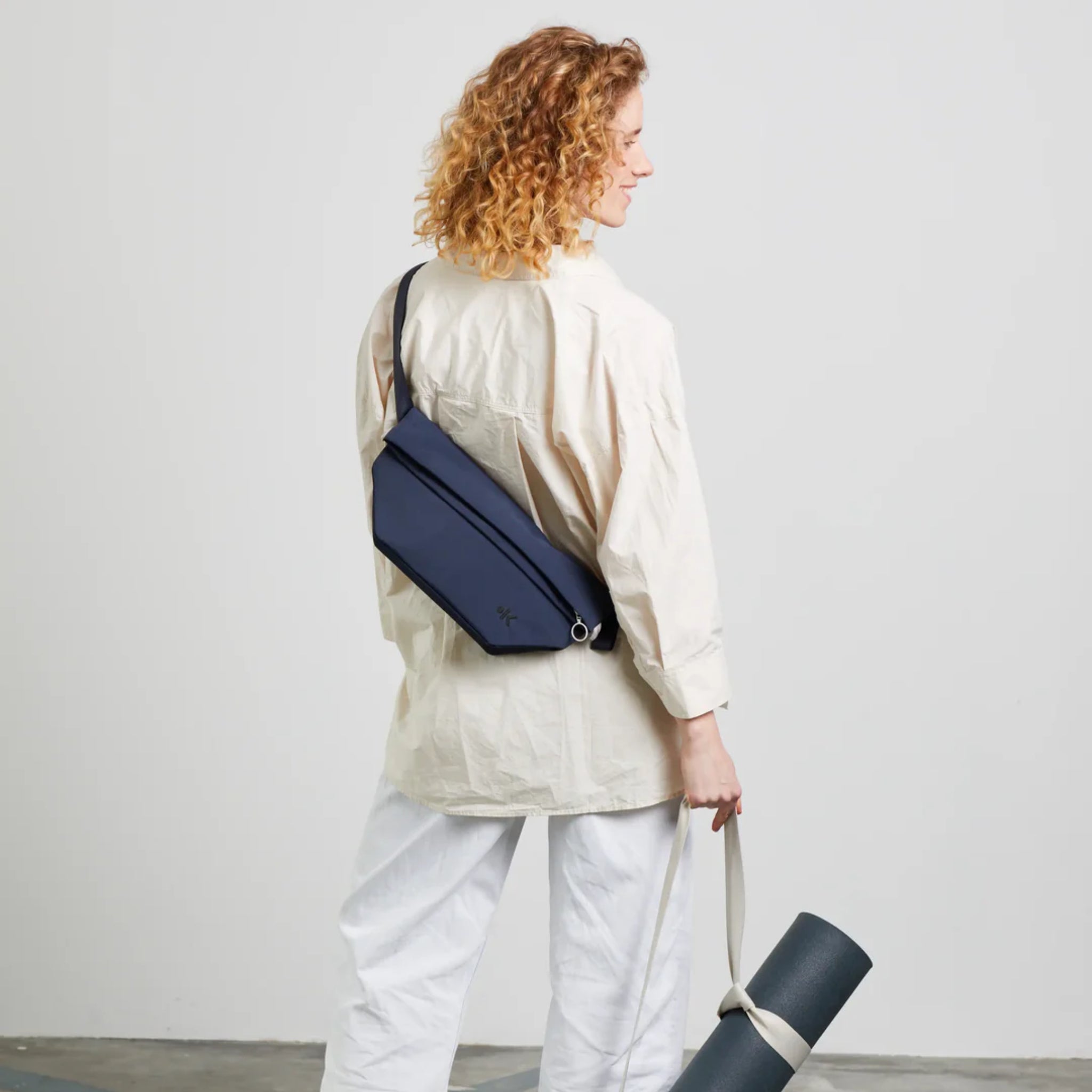 Eine Person mit lockigem Haar, die ein lockeres cremefarbenes Hemd und eine weiße Hose trägt, steht mit dem Gesicht nach hinten, trägt eine dunkelblaue Umhängetasche und hält eine zusammengerollte Yogamatte.