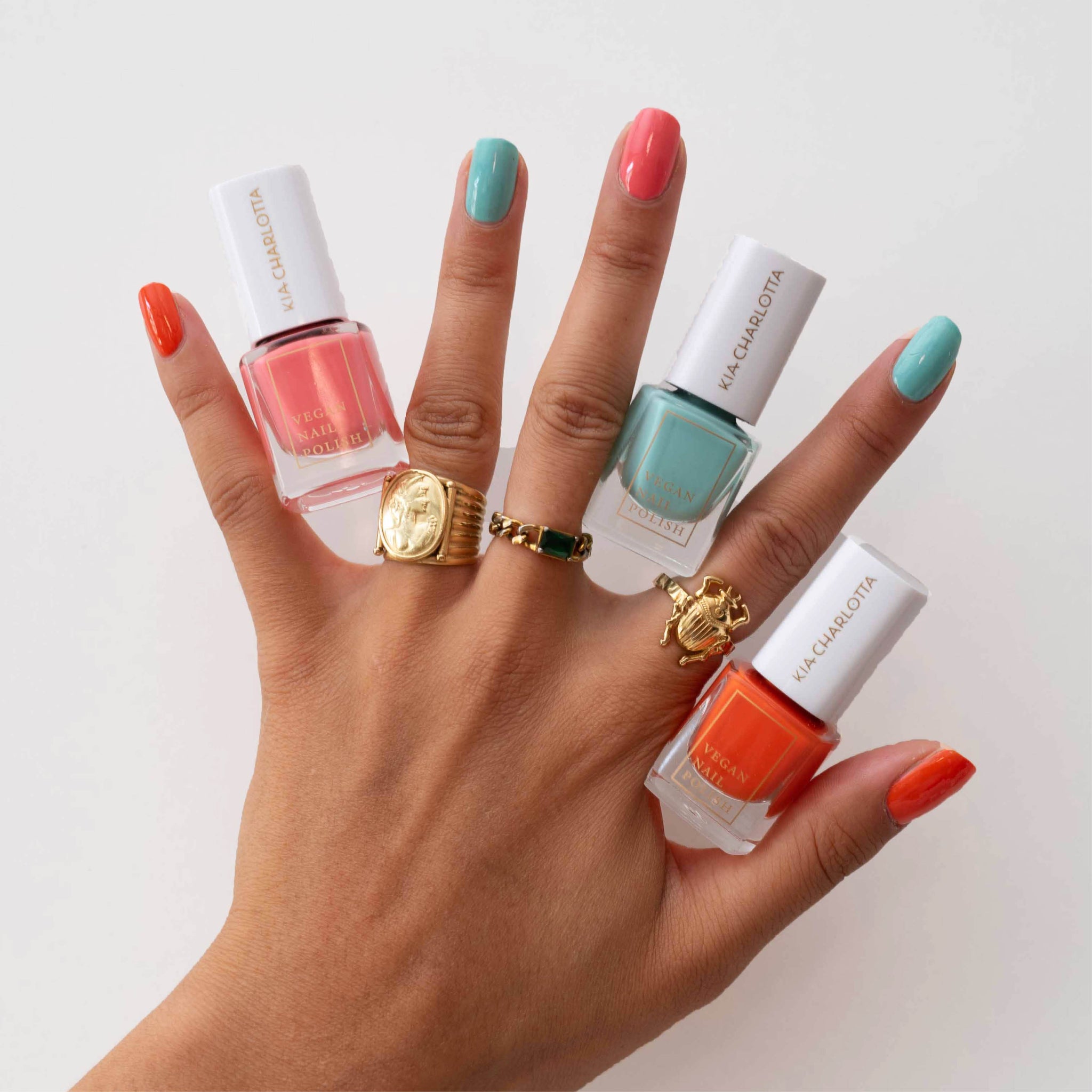 Hand mit farbigen Nagellacken und goldenen Ringen, die Flaschen mit veganem Nagellack halten.