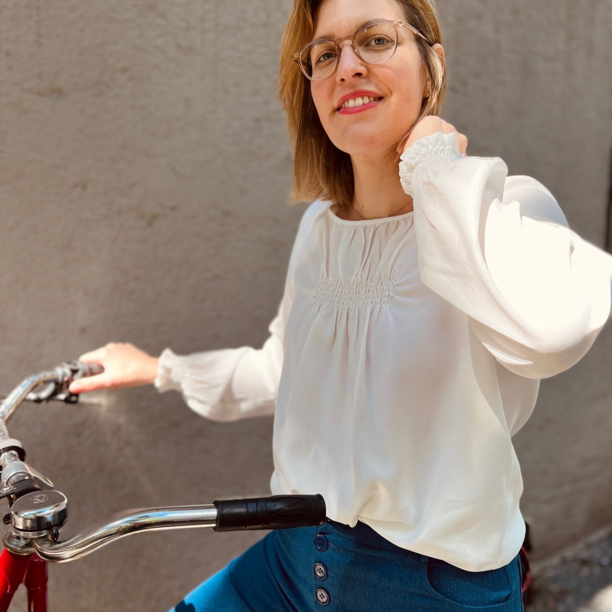 Frau mit Brille lächelt in einer weißen Bluse, sitzt auf einem Fahrrad vor einer grauen Wand.