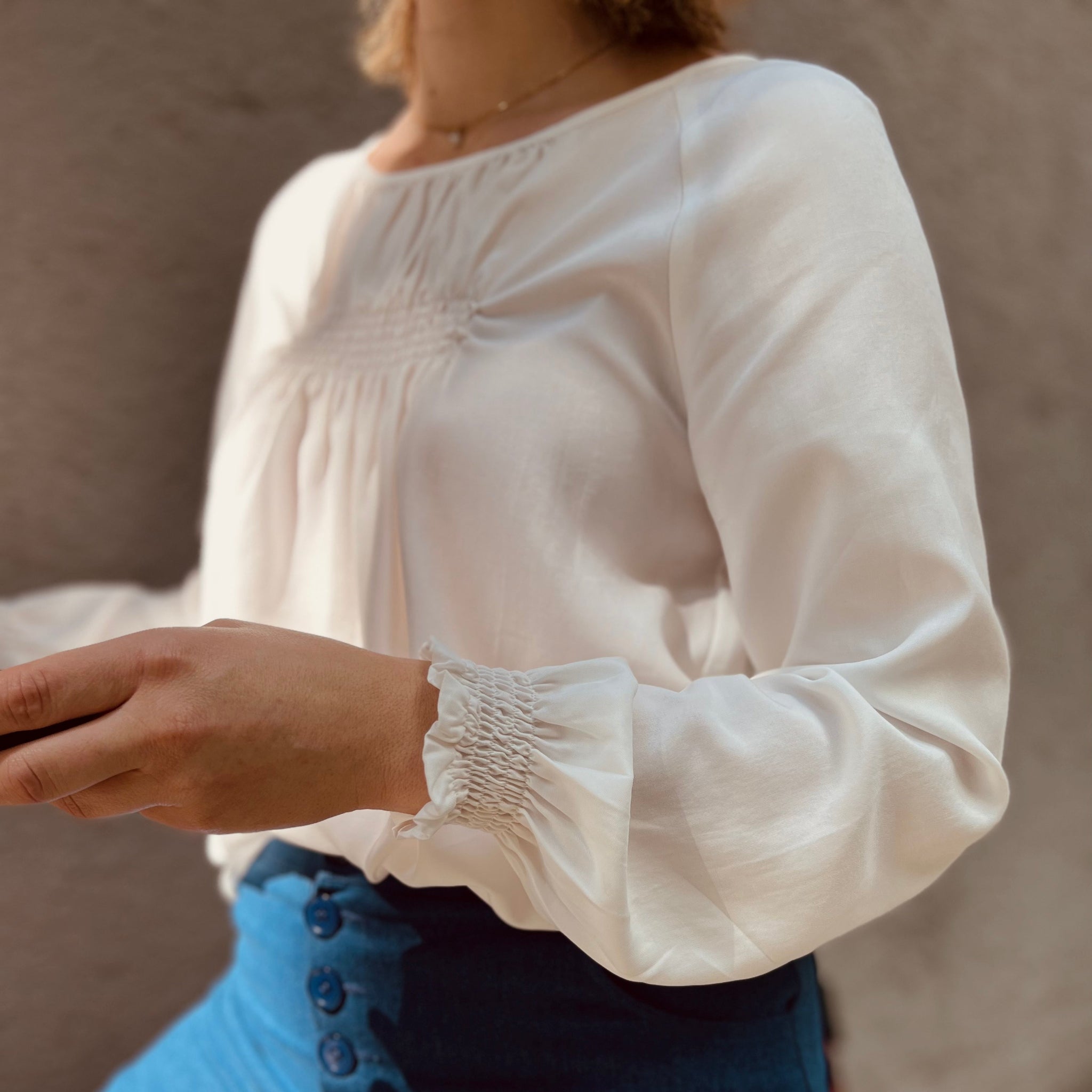 Frau in weißer Bluse und blauer Hose mit Knöpfen, vor neutralem Hintergrund.