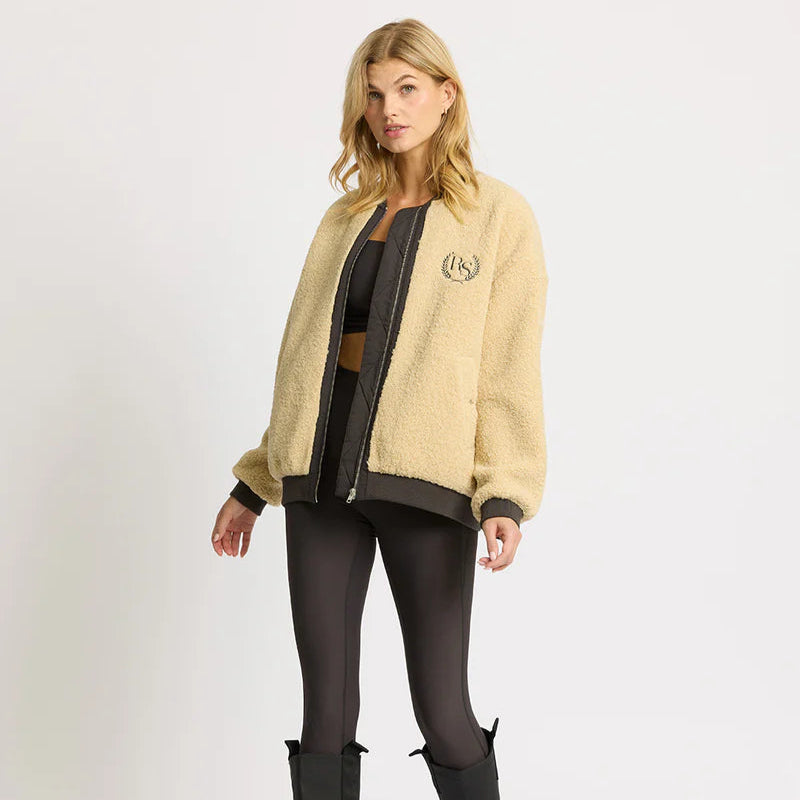 Eine Frau steht vor einem schlichten Hintergrund und trägt eine hellbraune Fleece-Jacke über einem schwarzen Sport-BH, schwarze Leggings mit hoher Taille und schwarze Stiefel. Sie hat blondes Haar und einen neutralen Gesichtsausdruck.