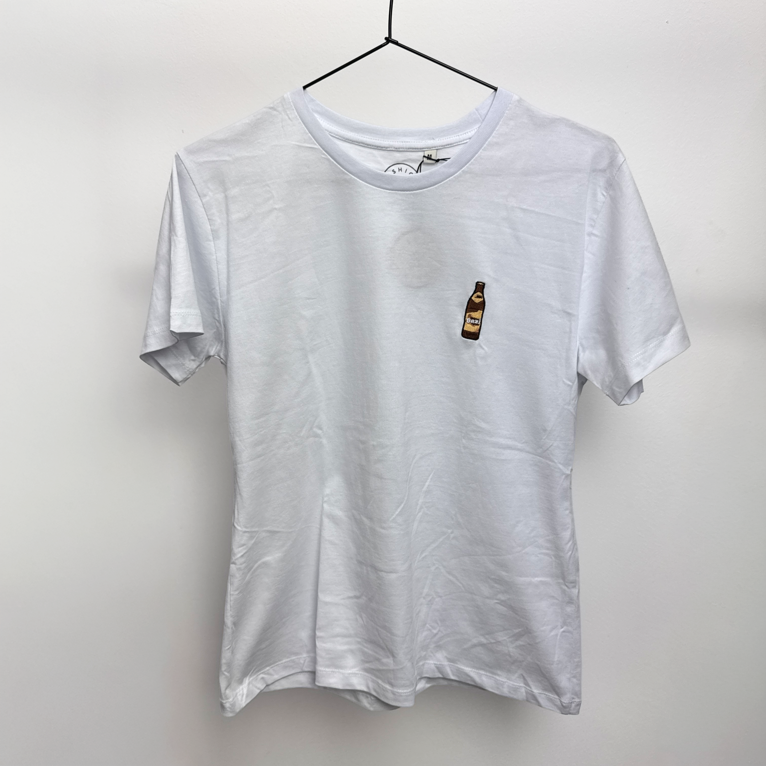 Ein schlichtes weißes T-Shirt auf einem Bügel mit einer kleinen gestickten Abbildung einer braunen Flasche auf der linken Brustseite.