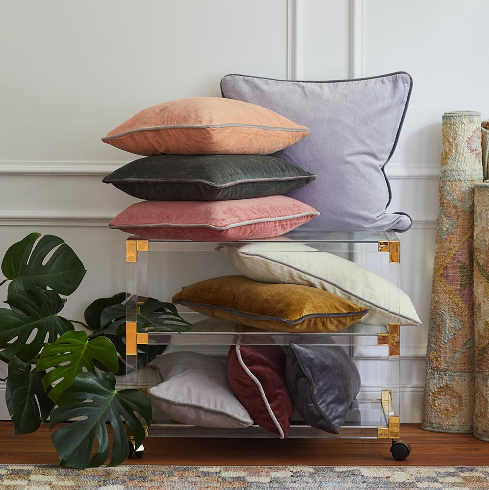 Ein Stapel bunter Samtkissen liegt auf einem durchsichtigen Acrylwagen mit goldenen Akzenten neben einer großen grünen Monstera-Pflanze und gerollten Teppichen, vor einer weiß getäfelten Wand und einem gemusterten Teppich.