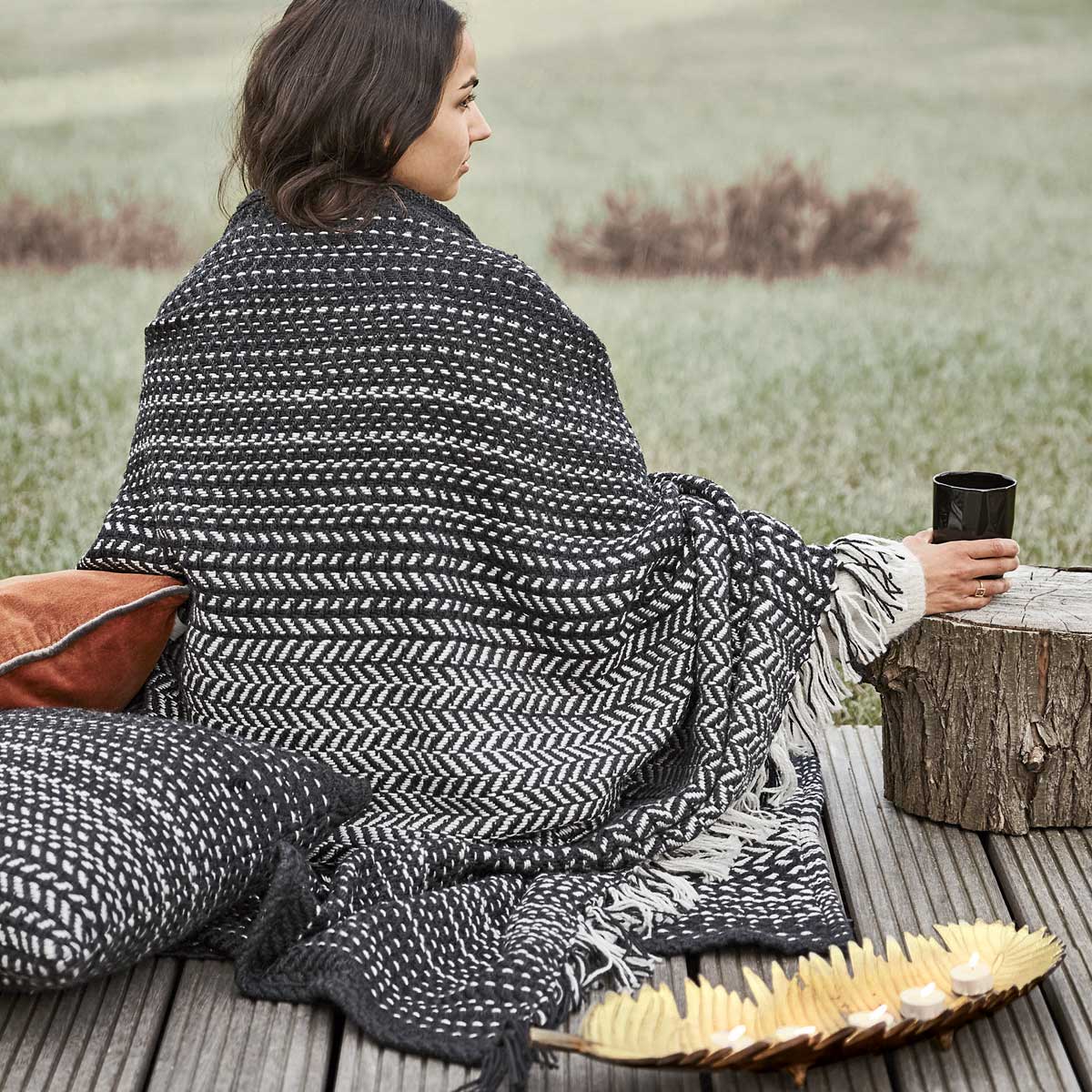 Eine in eine schwarz-weiß gemusterte Decke gehüllte Frau sitzt auf einer Holzterrasse und hält einen schwarzen Becher in der Hand. In der Nähe befinden sich Kissen und ein Teller mit Snacks, im Hintergrund sind Gras und Pflanzen zu sehen.