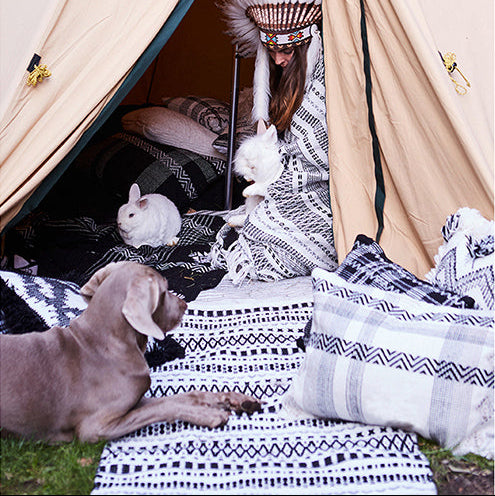 Ein Kind mit gefiedertem Kopfschmuck sitzt in einem beigen Zelt mit zwei weißen Kaninchen. Ein grauer Hund liegt außerhalb des Zeltes auf gemusterten Decken und Kissen. Die Szene spielt im Freien auf einer Wiese.
