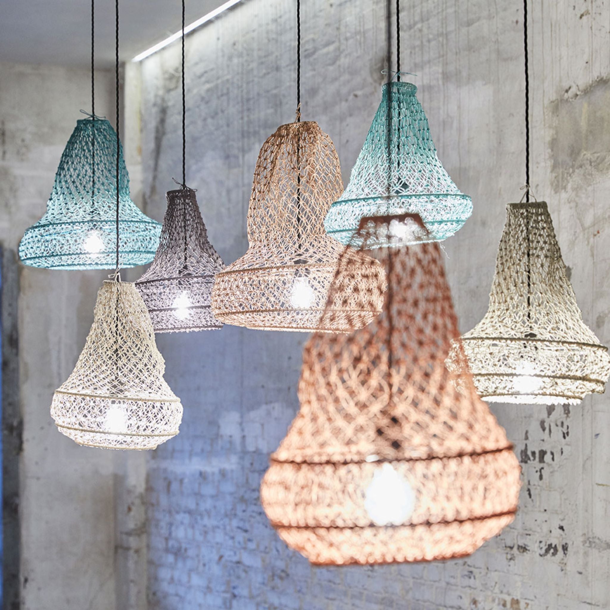 Eine Gruppe von Weidengeflecht-Hängelampenschirmen in verschiedenen Pastellfarben hängt von der Decke und beleuchtet eine rustikale, weiß gekalkte Ziegelwand im Hintergrund.
