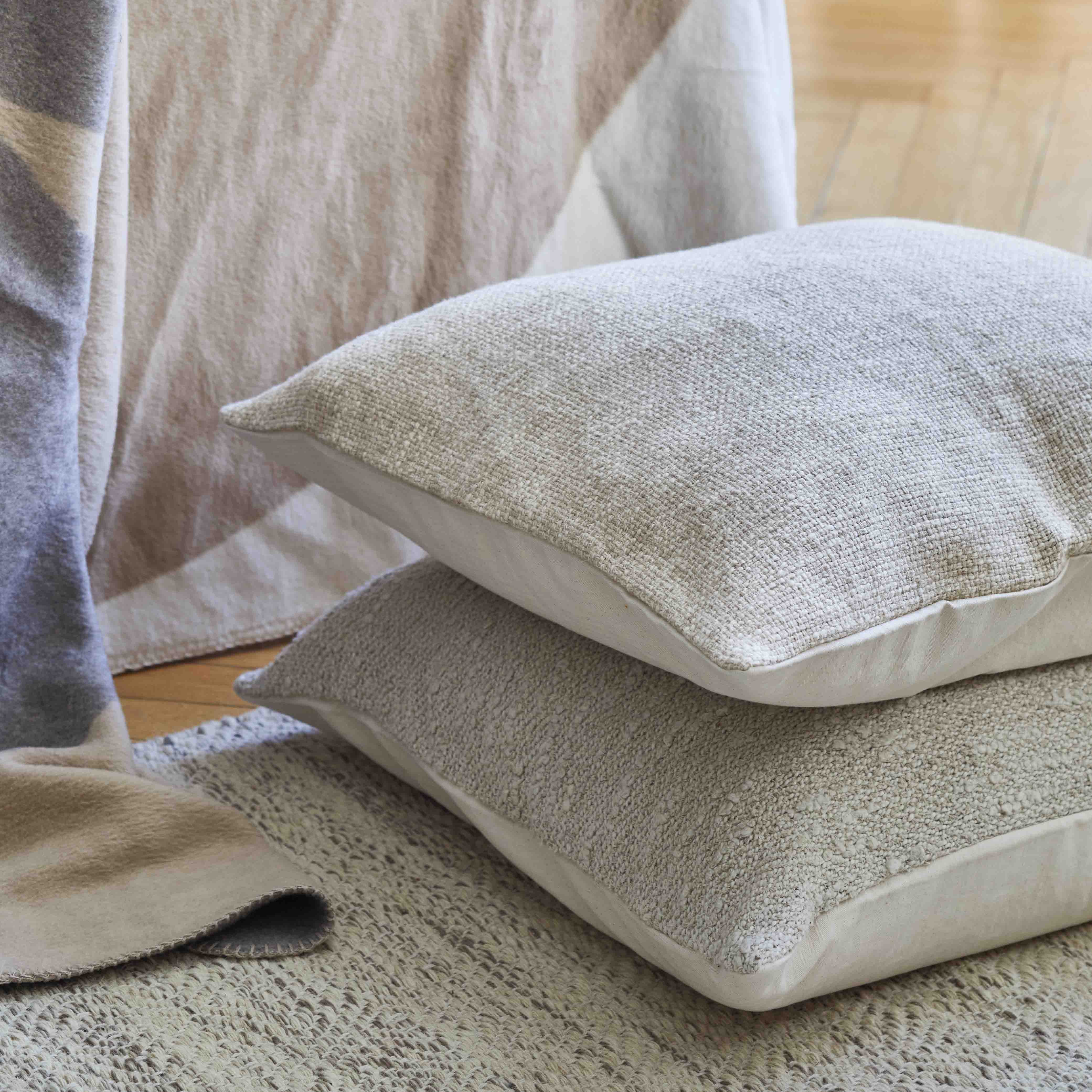Zwei beigefarbene, strukturierte Kissen liegen auf einem neutralen Webteppich, daneben ist eine weiche, gemusterte Decke auf einem Holzboden drapiert. Die Umgebung vermittelt eine gemütliche, minimalistische und ruhige Atmosphäre.