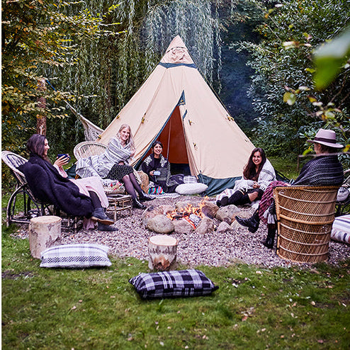 Fünf Personen sitzen um ein Lagerfeuer vor einem großen beigen Zelt in einem Waldgebiet, umgeben von Bäumen und Kissen, und genießen ein gemütliches Beisammensein im Freien.