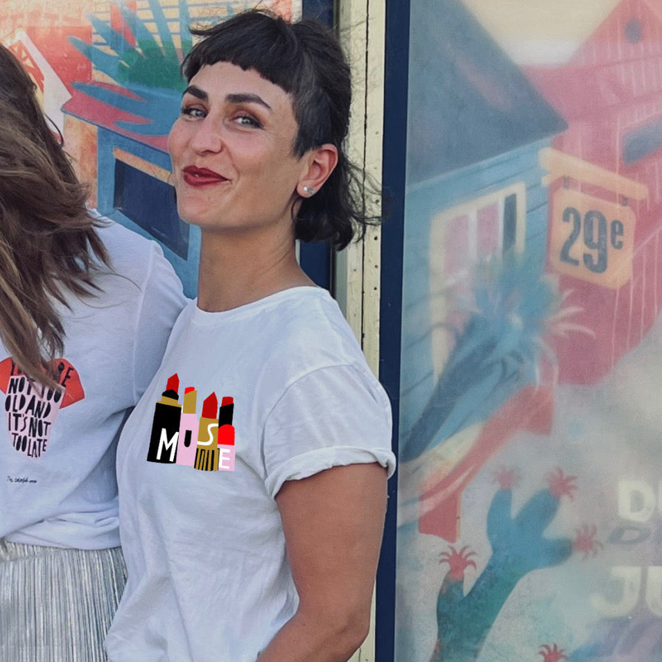 Eine Frau mit kurzen dunklen Haaren und rotem Lippenstift lächelt in die Kamera und trägt ein weißes T-Shirt mit bunten Motiven. Eine weitere Person steht neben ihr, meist außerhalb des Bildausschnitts. Im Hintergrund ist eine bunte, künstlerische Wandmalerei zu sehen.