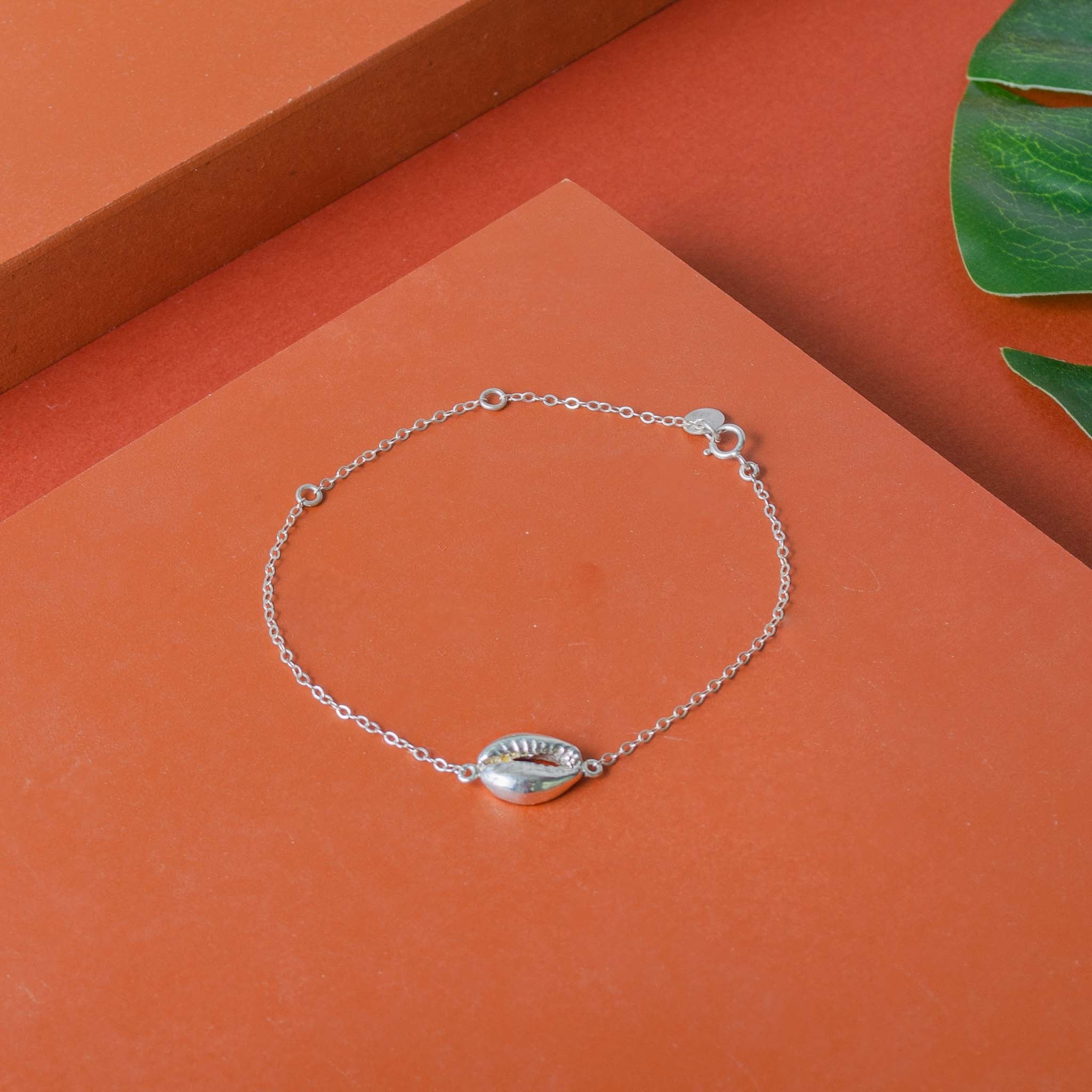 Ein zartes Silberarmband mit einem Kaurimuschel-Charm auf einer orangefarbenen Oberfläche, mit grünen Blättern in der oberen rechten Ecke.