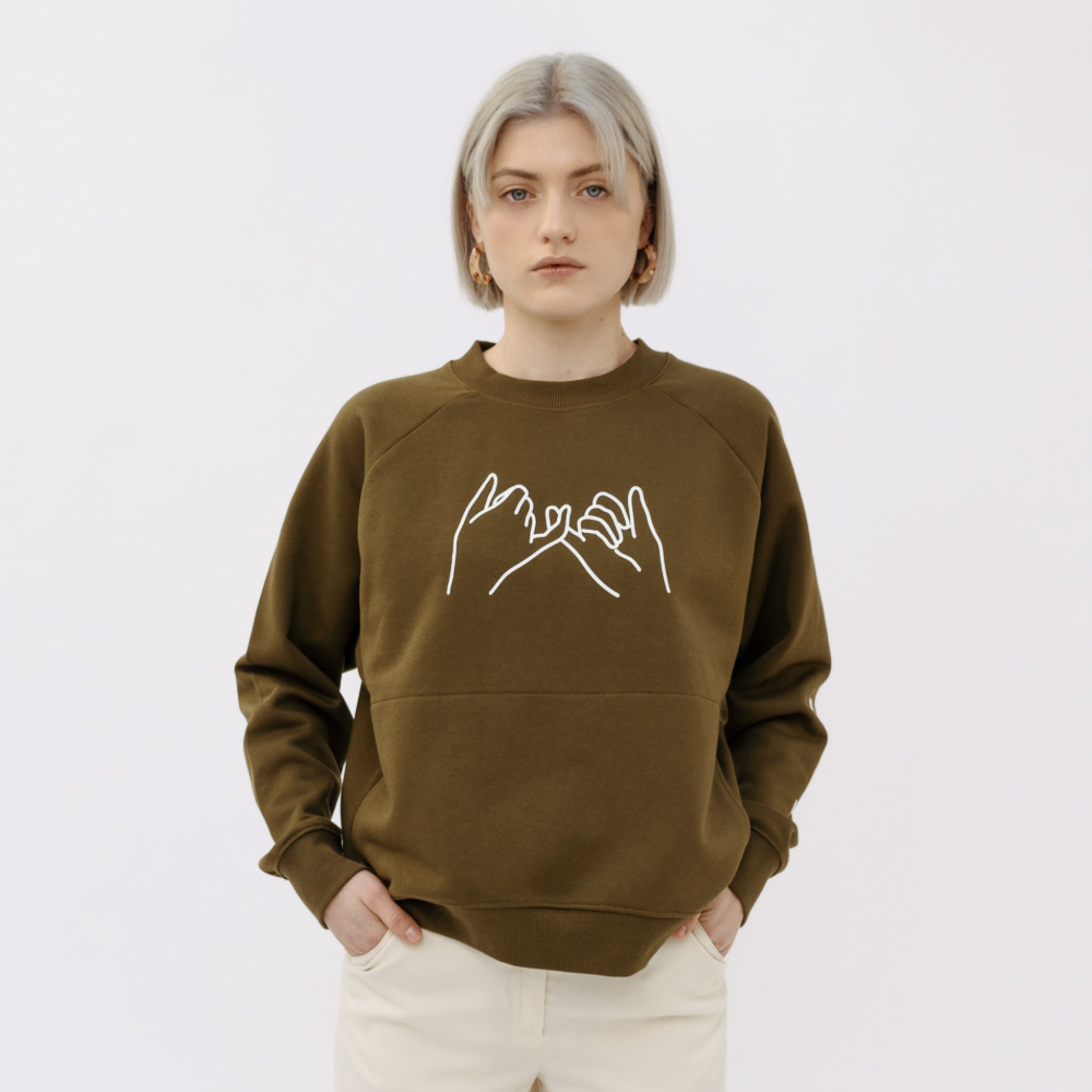 Frau mit blonden Haaren trägt einen olivgrünen Pullover mit einem weißen Aufdruck von ineinander verschränkten Händen.