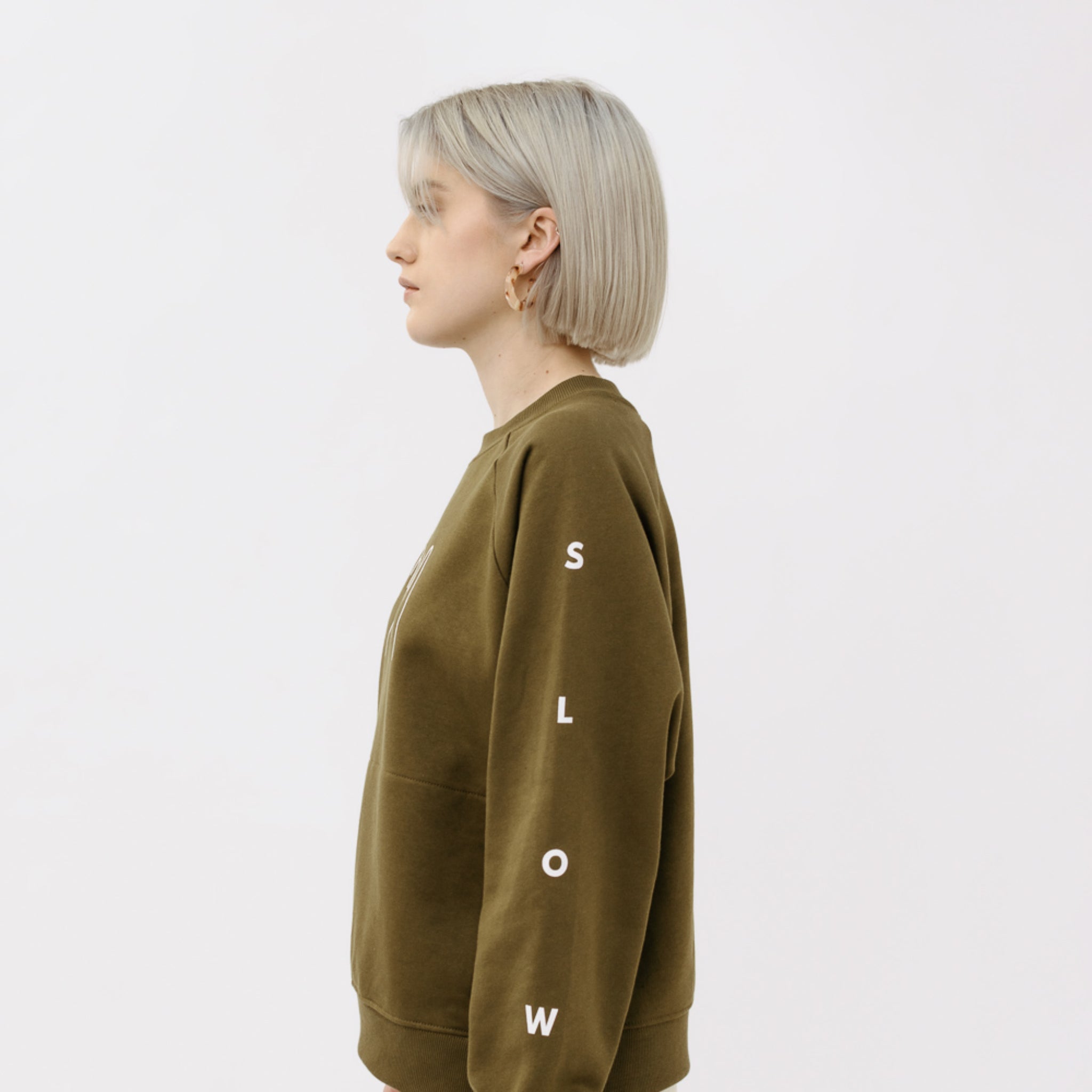 Person mit blondem Bob trägt einen olivgrünen Pullover, auf dem Ärmel steht "SLOW", auf einem neutralen Hintergrund.