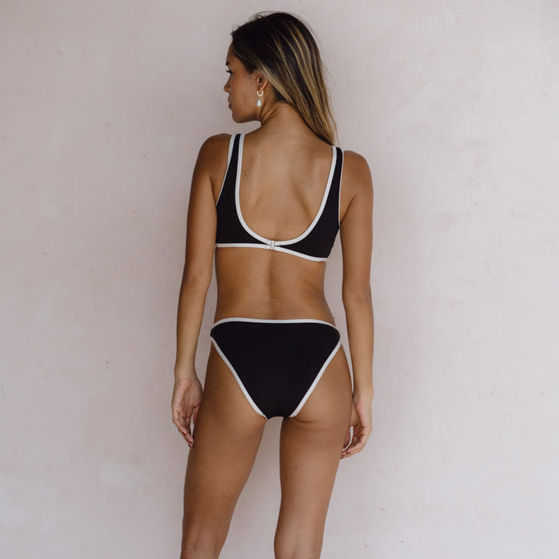 Eine Frau steht mit dem Rücken zur Kamera, trägt einen schwarzen Bikini mit weißem Besatz, Ohrringe und offenes Haar vor einer schlichten hellen Wand.