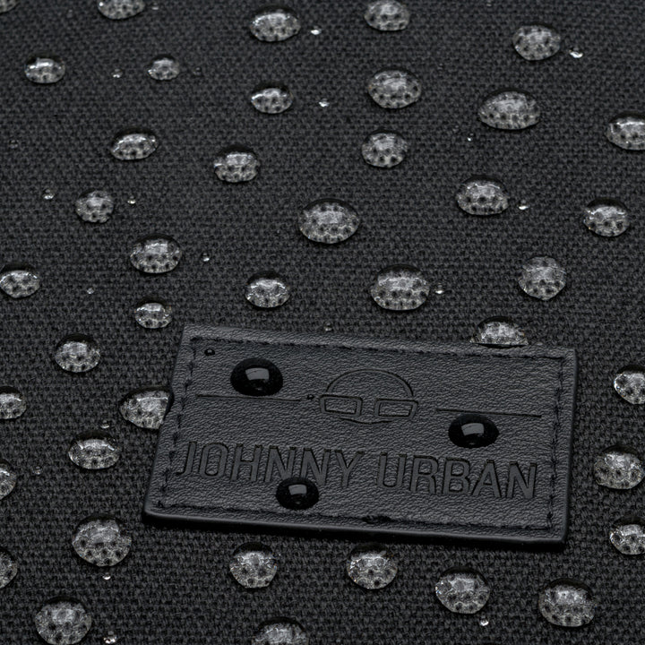 Nahaufnahme eines schwarzen Stoffes mit erhabenen, glänzenden Punktmustern und einem rechteckigen, schwarz genähten Etikett mit der Aufschrift JOHNNY URBAN und einer kleinen Grafik über dem Text.