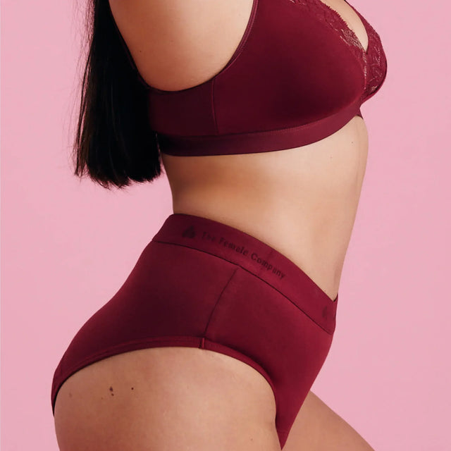 Eine Frau, die einen passenden weinroten BH und hochtaillierte Unterwäsche trägt, posiert vor einem rosa Hintergrund und zeigt eine Seitenansicht ihres Körpers von den Schultern bis zu den Oberschenkeln.