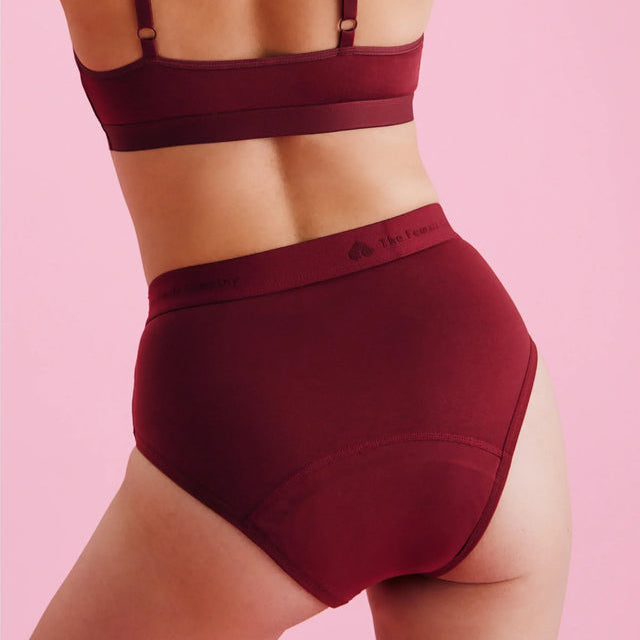 Eine Person, die einen passenden bordeauxroten BH und hochtaillierte Unterwäsche trägt, steht vor einem rosafarbenen Hintergrund und zeigt ihren Rücken und Unterkörper.