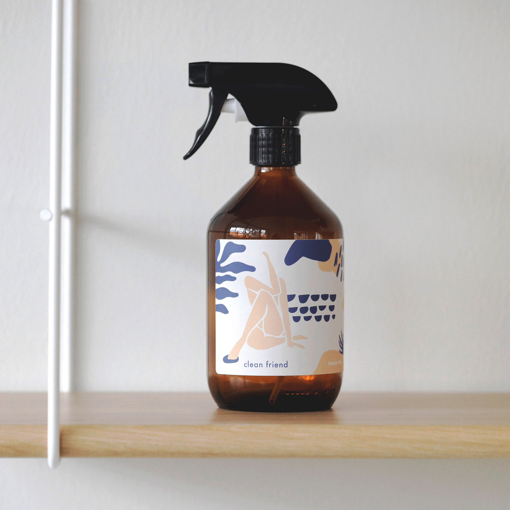 Braune Sprühflasche mit kunstvollem Etikett steht auf einem Holzregal vor einer weißen Wand.