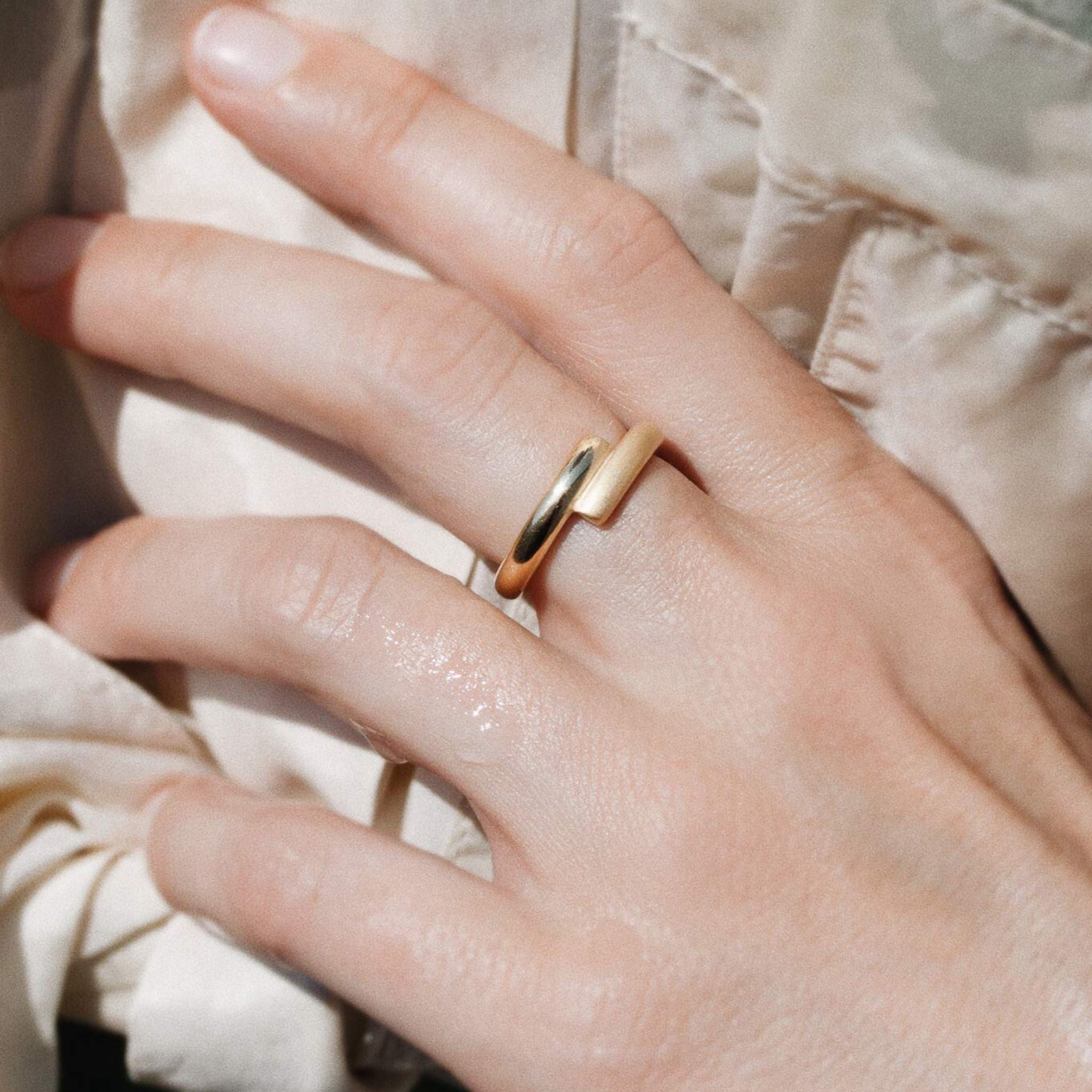 Nahaufnahme einer Hand, die einen modernen, minimalistischen Goldring am Mittelfinger trägt. Der Hintergrund ist ein heller, strukturierter Stoff, und die Beleuchtung hebt die Glätte der Haut und des Rings hervor.
