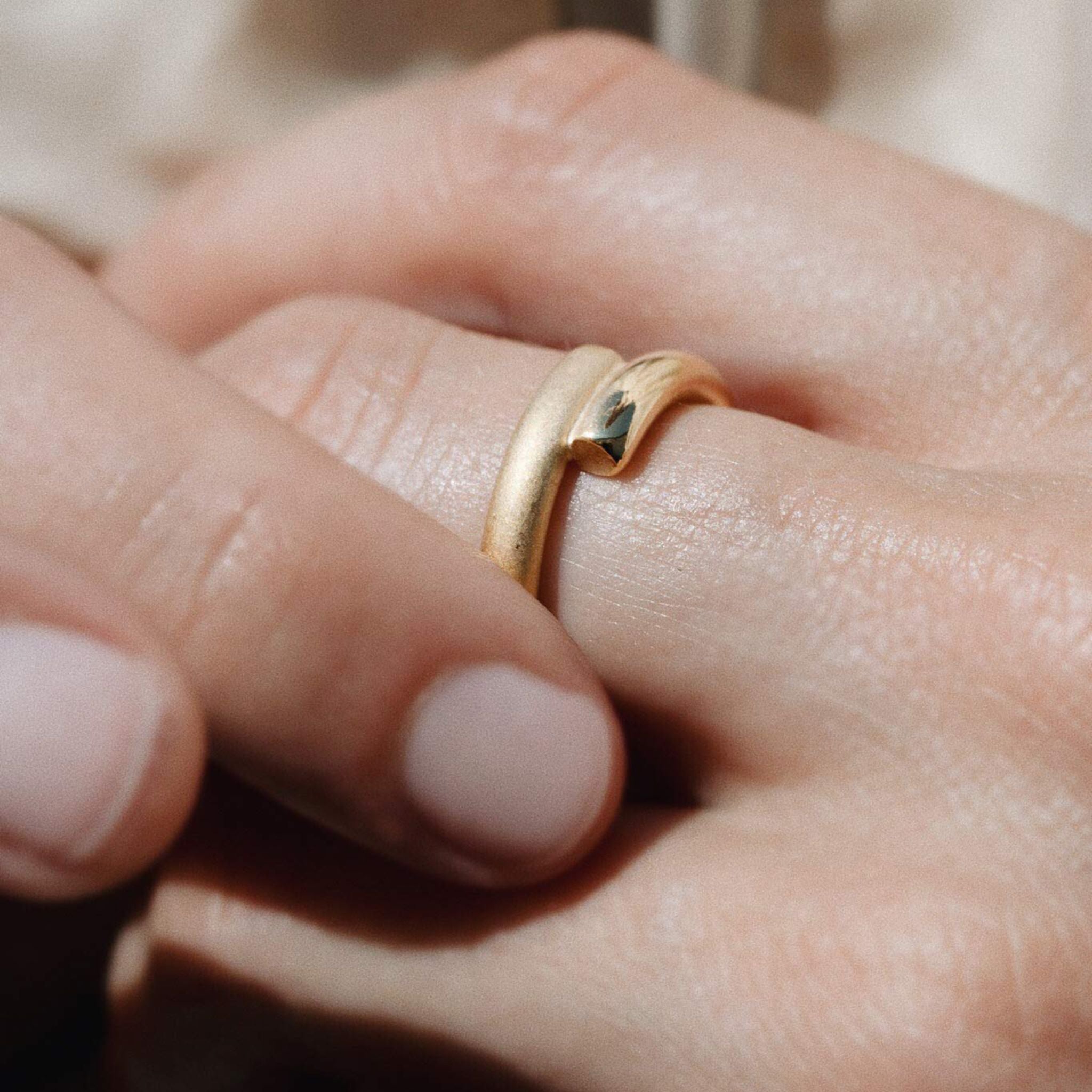 Nahaufnahme einer Hand, die einen mattgoldenen Ring mit einem dezenten Wellenmuster am Ringfinger trägt. Die Finger sind leicht gekrümmt, und die Nägel sind kurz und ordentlich manikürt.