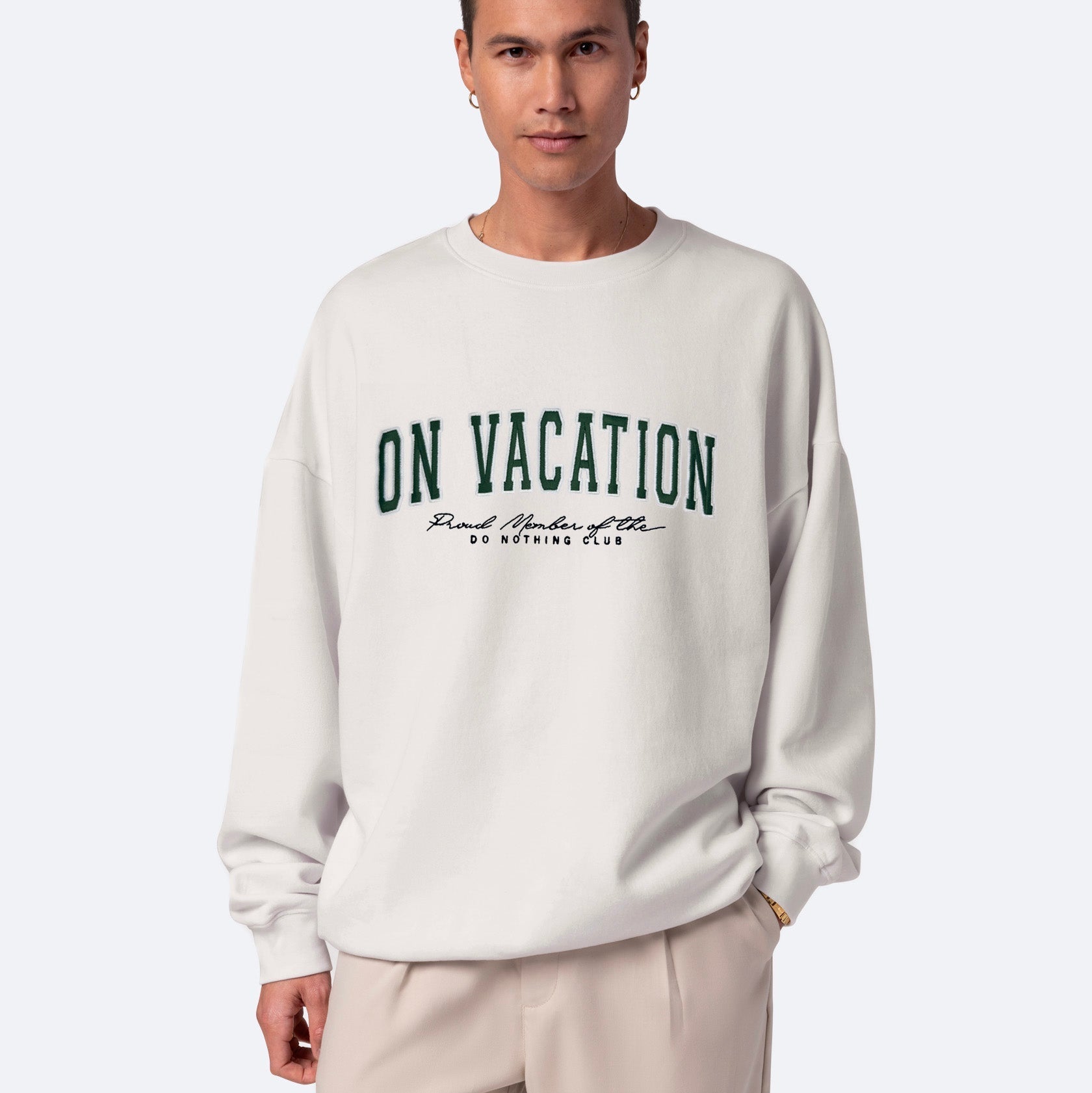 Eine Person trägt ein helles Sweatshirt mit grüner Aufschrift ON VACATION und darunter kleinerer schwarzer Aufschrift Good Minds Club DO NOTHING CLUB. Die Person hat kurze Haare und steht vor einem einfarbigen Hintergrund.