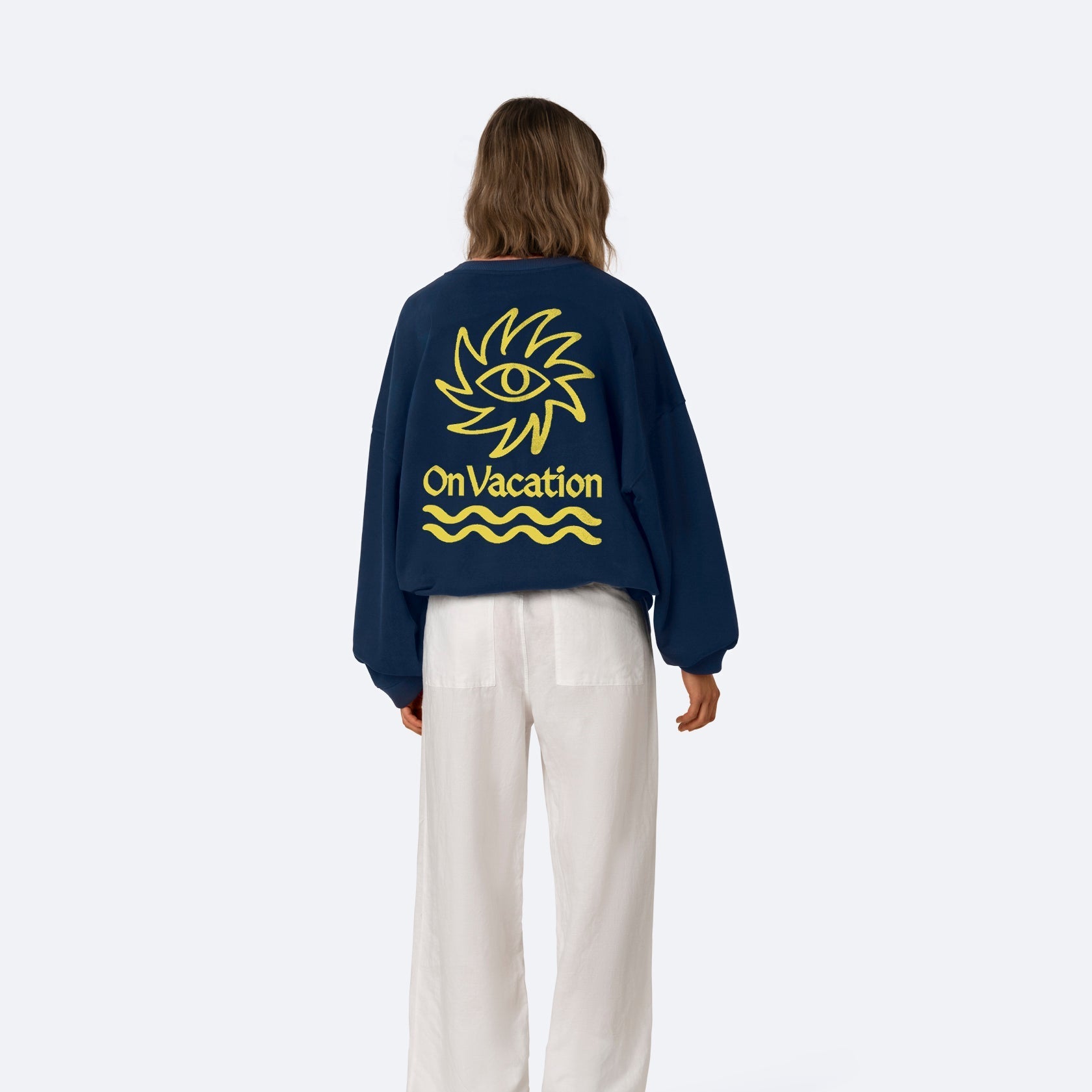 Eine Person steht mit dem Gesicht zur Wand, trägt ein marineblaues Sweatshirt mit einer gelben Sonnen- und Augengrafik und der Aufschrift On Vacation auf dem Rücken, dazu eine weite weiße Hose und beigefarbene Schuhe.