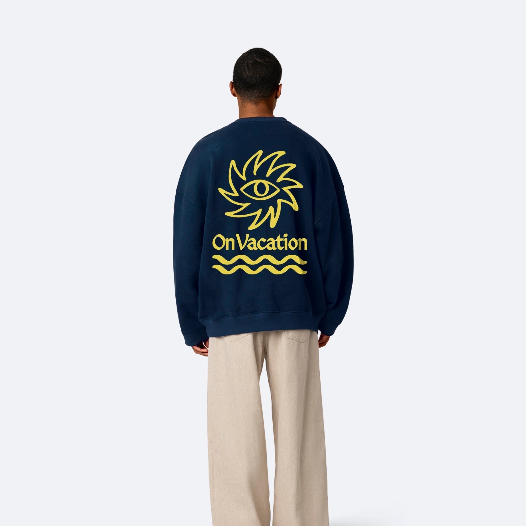 Eine Person steht mit dem Gesicht zur Wand und trägt ein marineblaues Sweatshirt mit einem großen gelben Auge in der Sonne und dem Schriftzug On Vacation über Wellenlinien, gepaart mit einer lockeren, hellbeigen Hose.