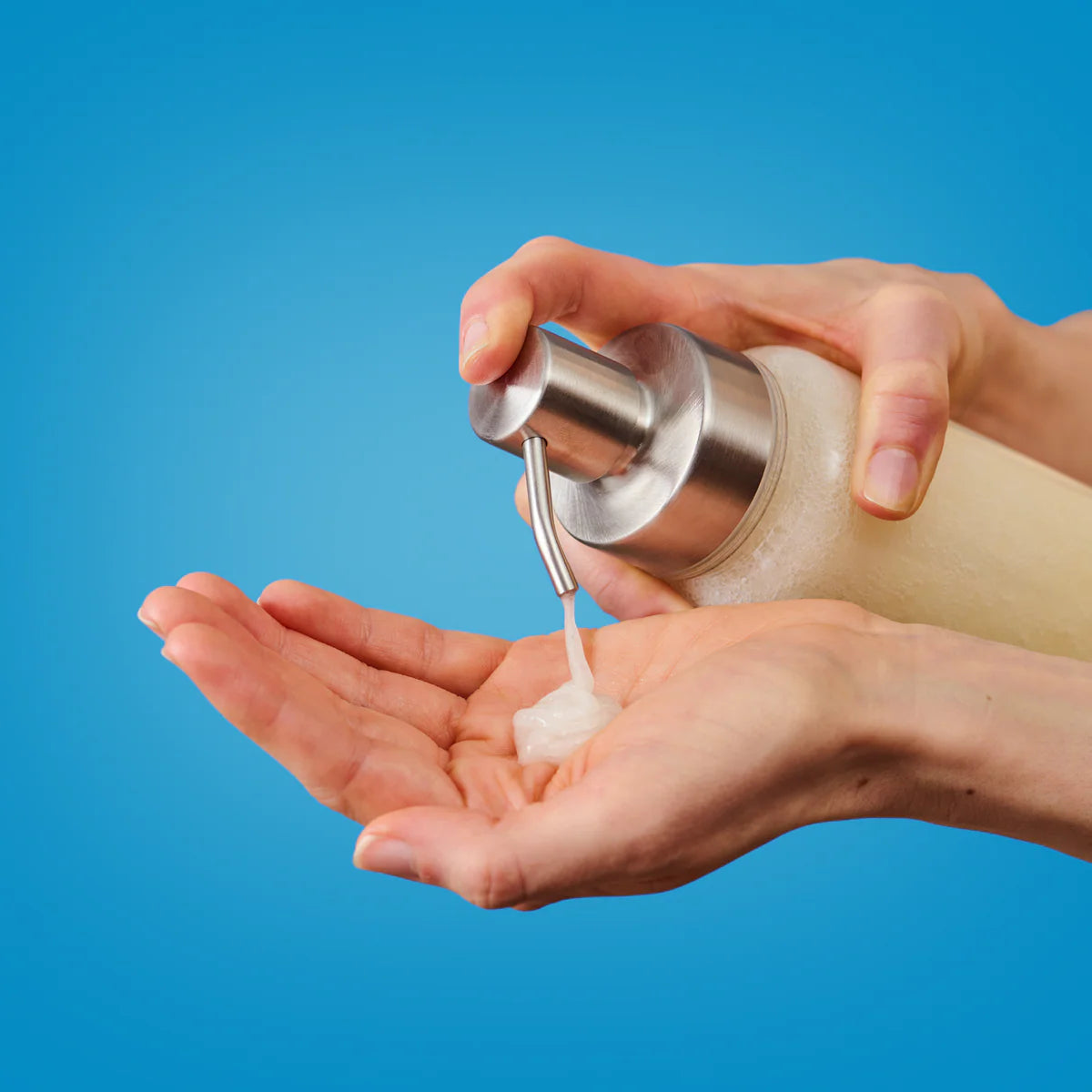 Eine Person gibt vor einem blauen Hintergrund eine cremige Flüssigkeit aus einer Pumpflasche auf ihre Handfläche ab.