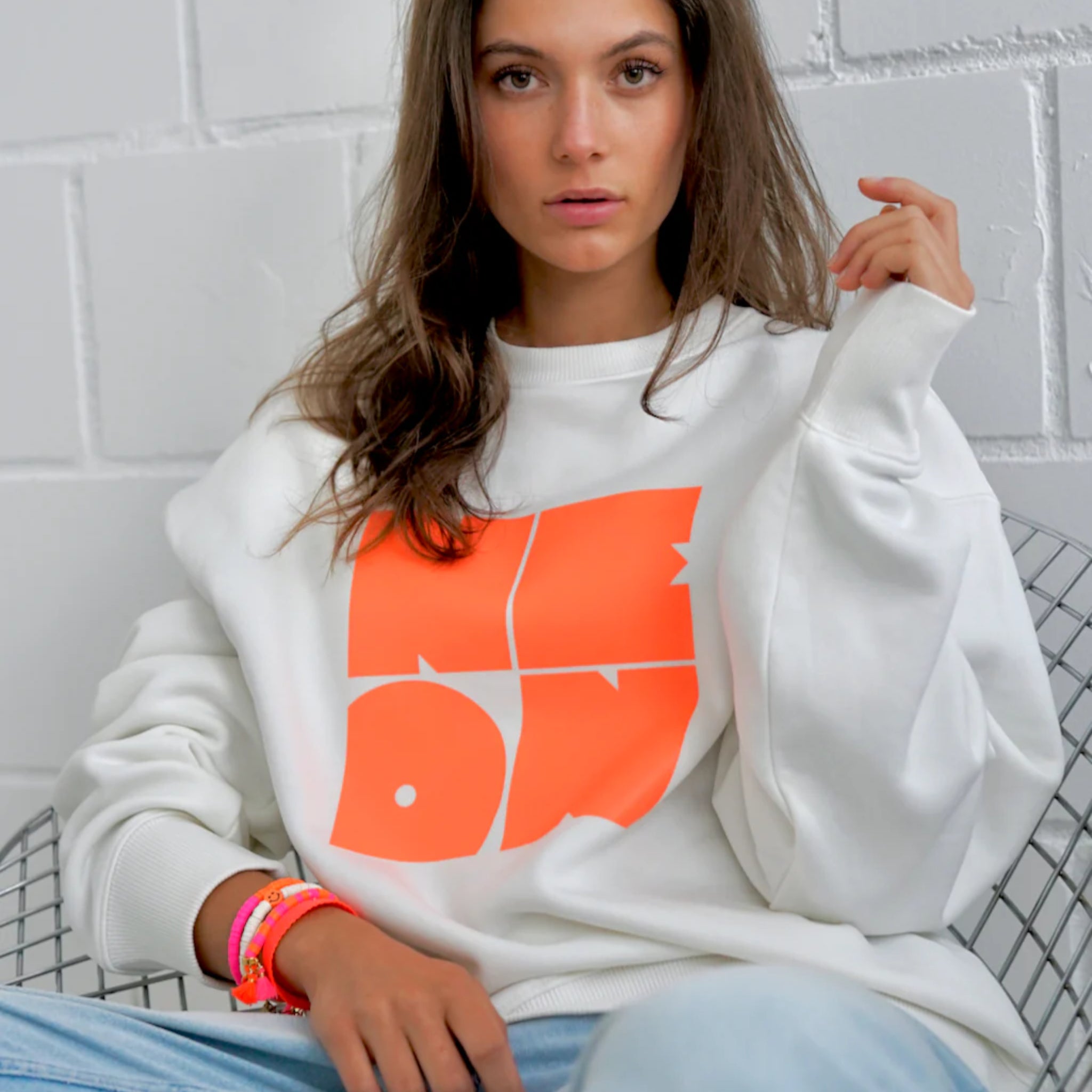 Eine Frau mit langen braunen Haaren sitzt auf einem Drahtstuhl und trägt ein weißes Sweatshirt mit großer oranger Schrift und bunten Armbändern. Sie posiert mit einer Hand, die ihr Haar berührt, gegen eine weiße Backsteinmauer.