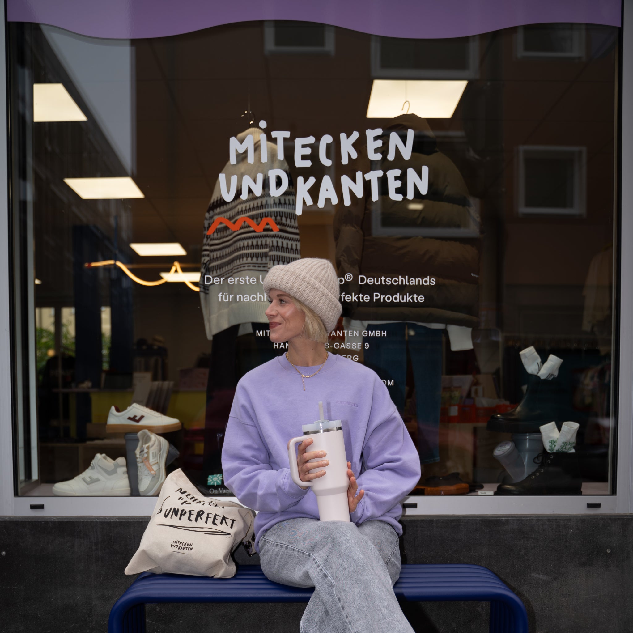 Eine Person sitzt mit einer großen Tasse vor einem Schaufenster mit der Aufschrift "Mitecken und Kanten".