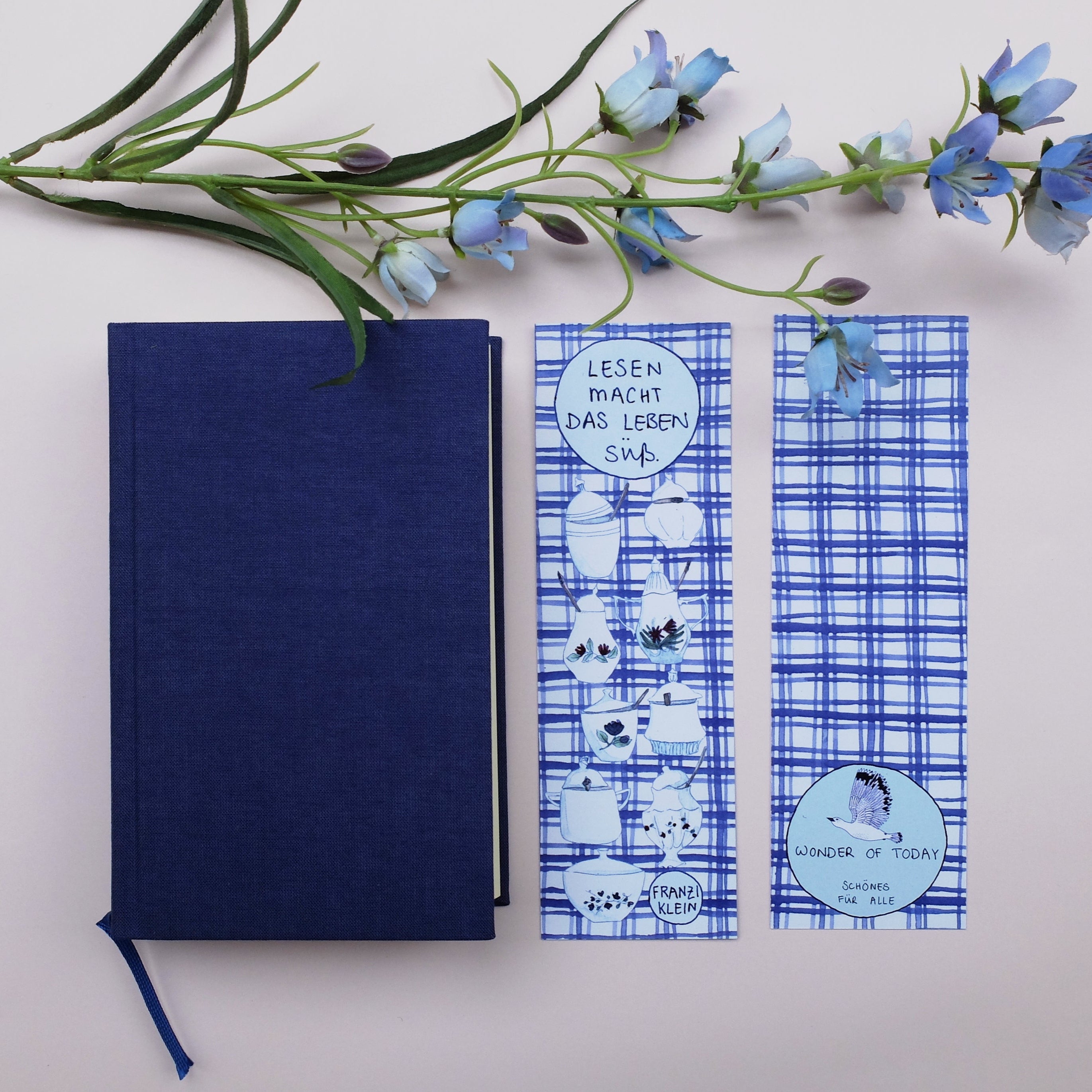 Ein blaues Buch mit festem Einband, zwei gemusterte blau-weiße Lesezeichen mit deutschem und englischem Text und ein Zweig mit blauen Blumen sind auf einem hellen Hintergrund angeordnet.