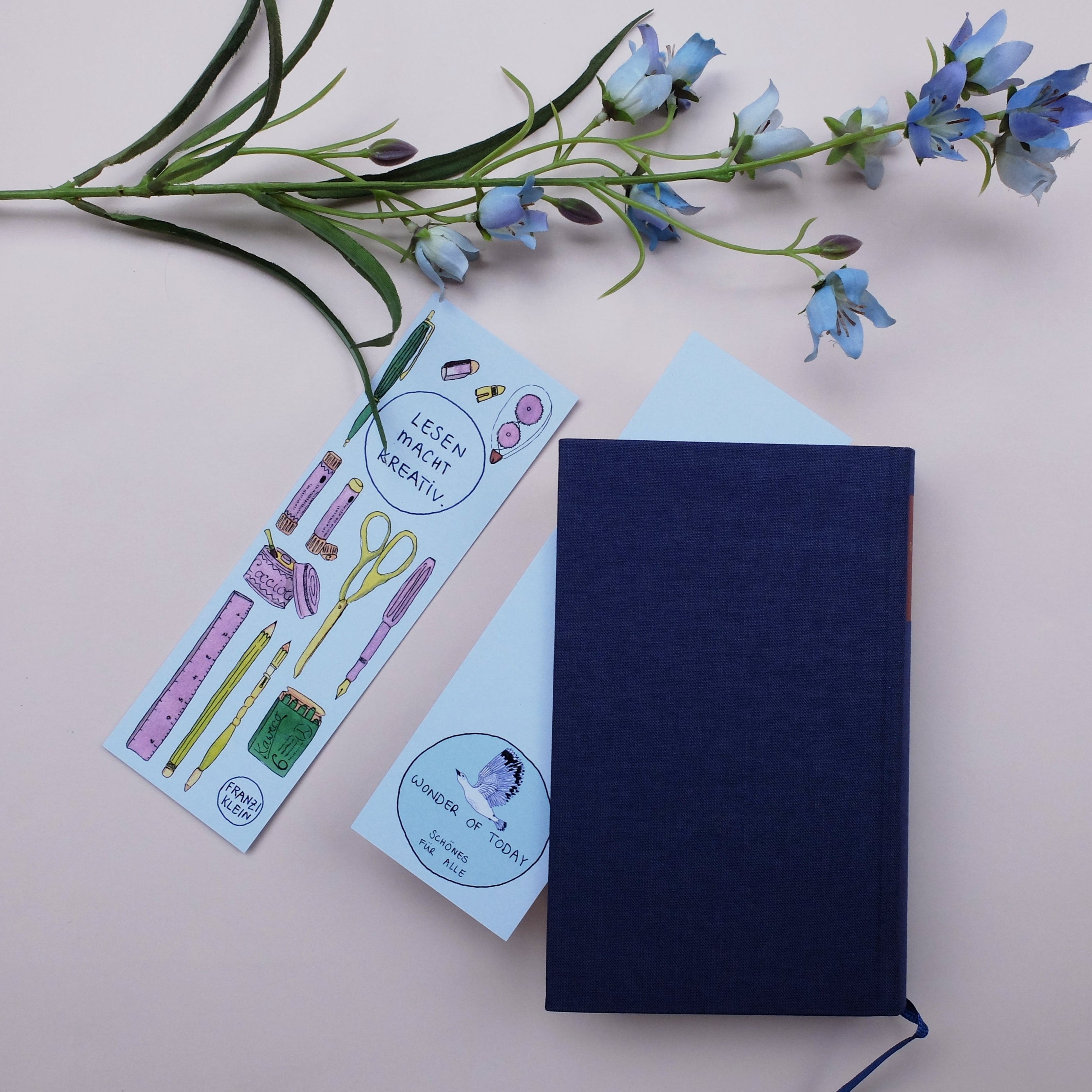 Ein dunkelblaues Notizbuch, ein hellblauer Umschlag, ein Lesezeichen im Schreibwarenstil und ein Zweig mit hellvioletten Blumen sind auf einer blassrosa Fläche angeordnet.