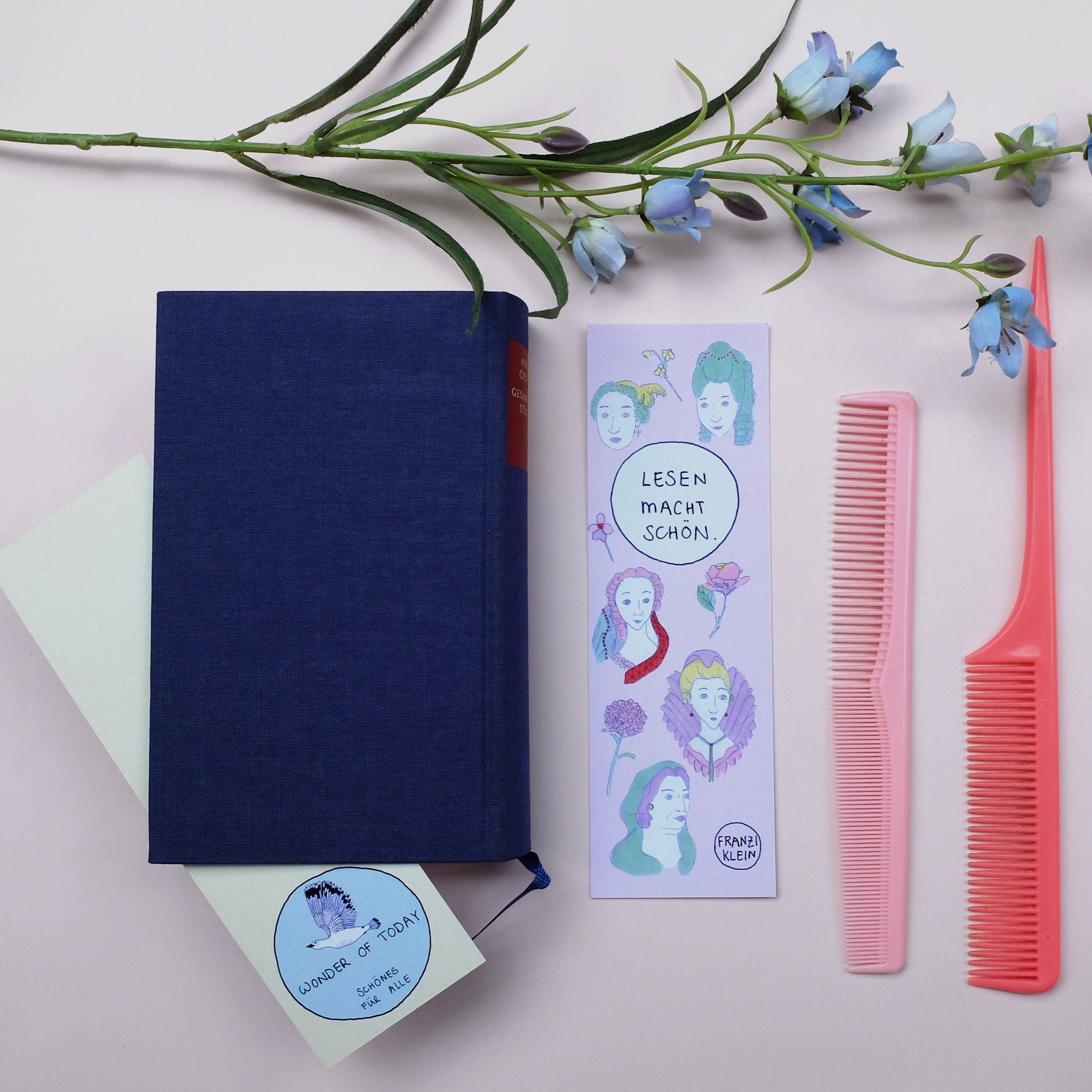 Ein marineblaues Buch, ein Lesezeichen mit illustrierten Gesichtern und deutschem Text, zwei rosa Kämme und blaue Blumen, die auf einer hellen Fläche angeordnet sind.