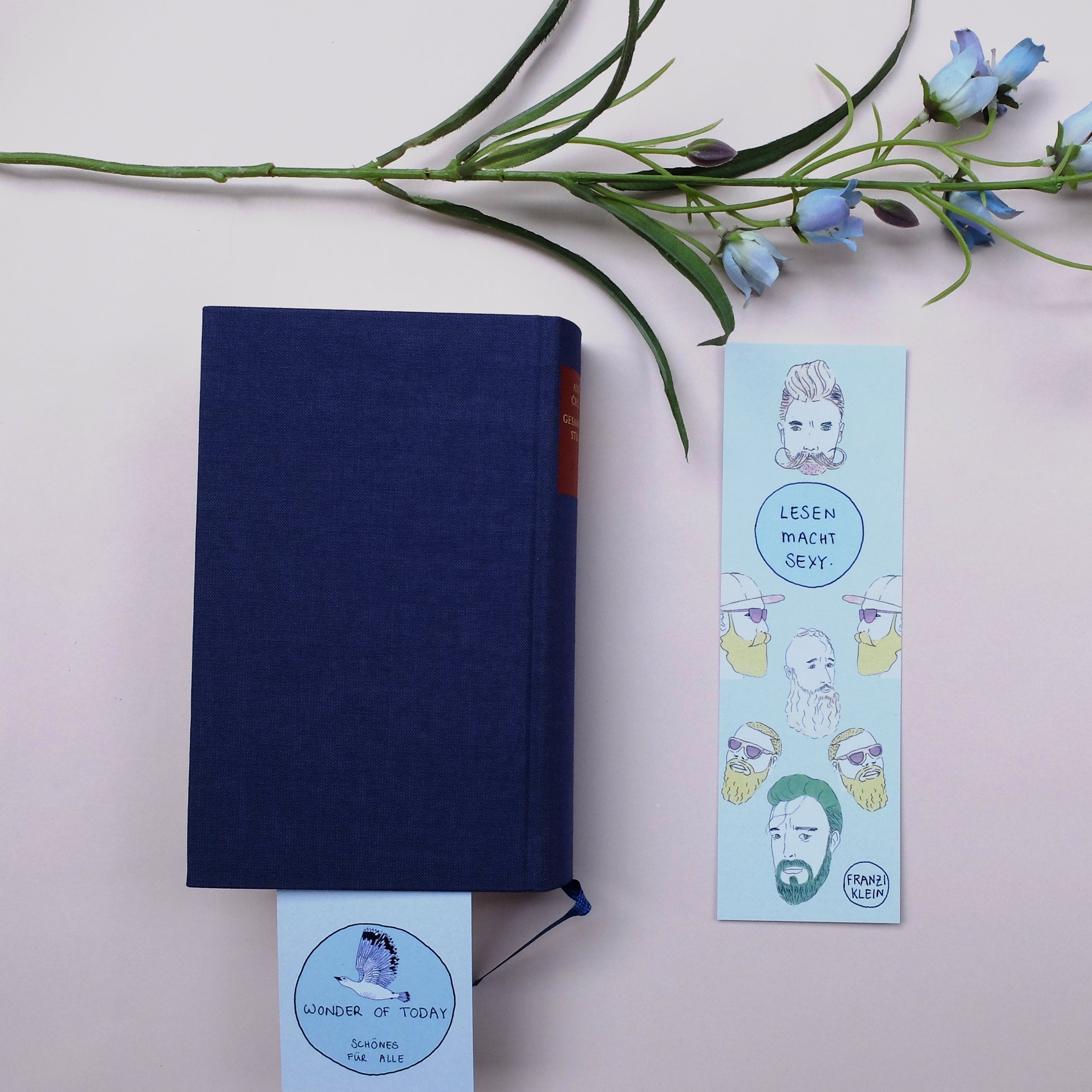 Ein geschlossenes blaues Buch mit einem hellblau illustrierten Lesezeichen, das sich teilweise darin befindet, liegt auf einer hellen Fläche neben einem anderen illustrierten Lesezeichen und einem Zweig mit lila Blumen.