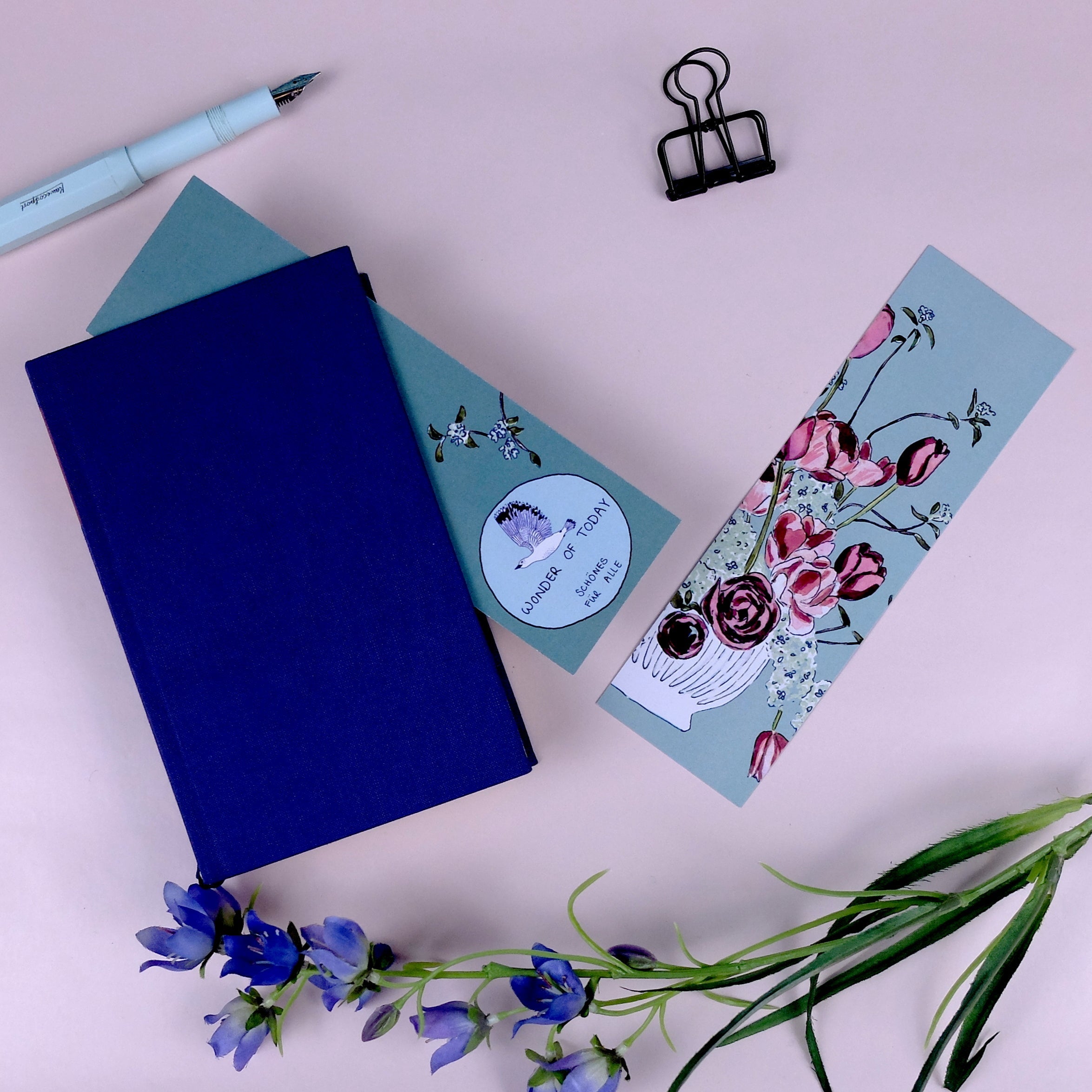 Ein blaues Notizbuch, ein grüner Umschlag mit einem Blumenaufkleber, ein geblümtes Lesezeichen, eine Heftklammer, ein Füllfederhalter und lila Blumen auf einem blassrosa Hintergrund.