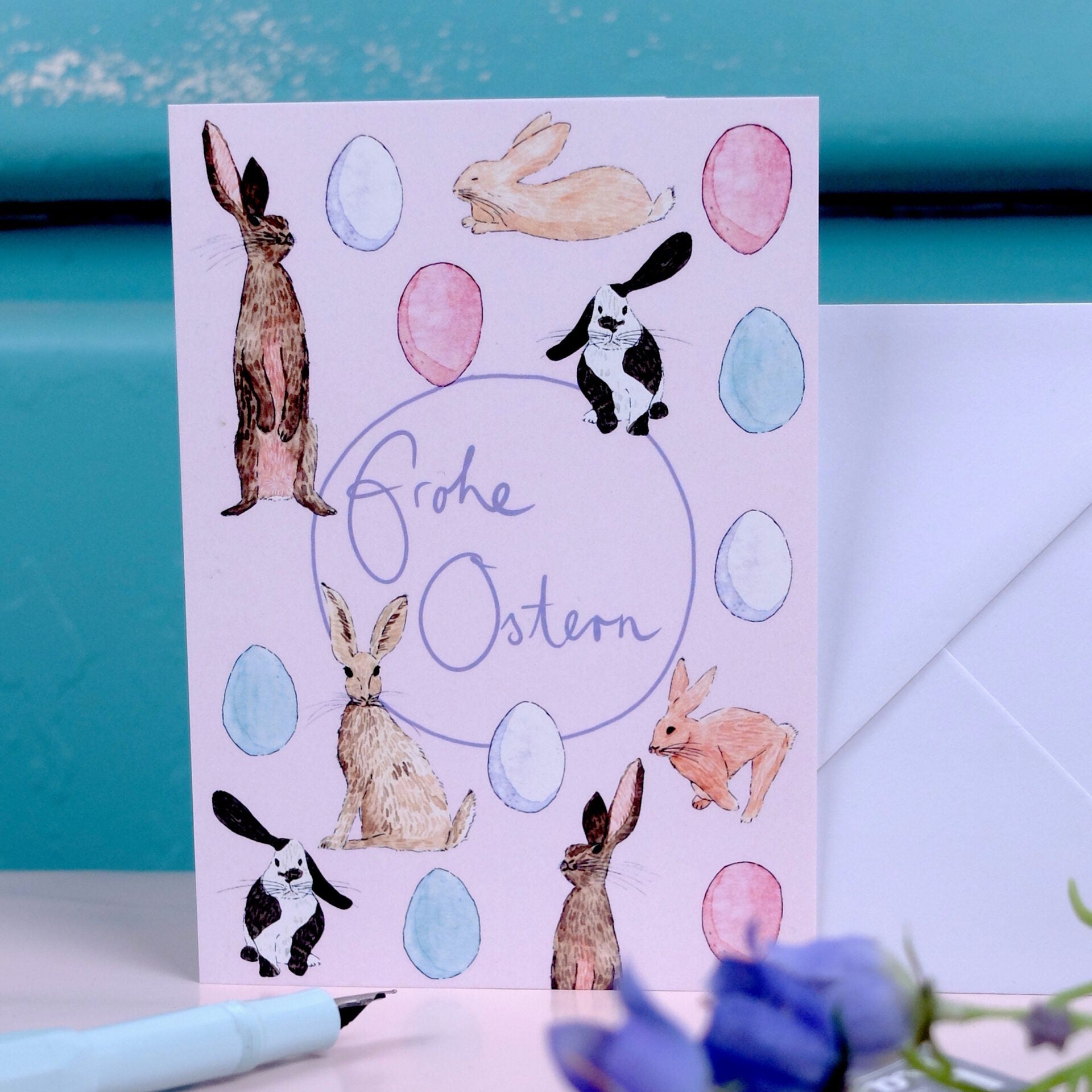 Eine pastellfarbene Osterkarte mit Illustrationen von Hasen und Eiern, mit dem handgeschriebenen deutschen Text Frohe Ostern in der Mitte. Ein Briefumschlag und ein Stift sind in der Nähe, mit verschwommenen Blumen im Vordergrund.