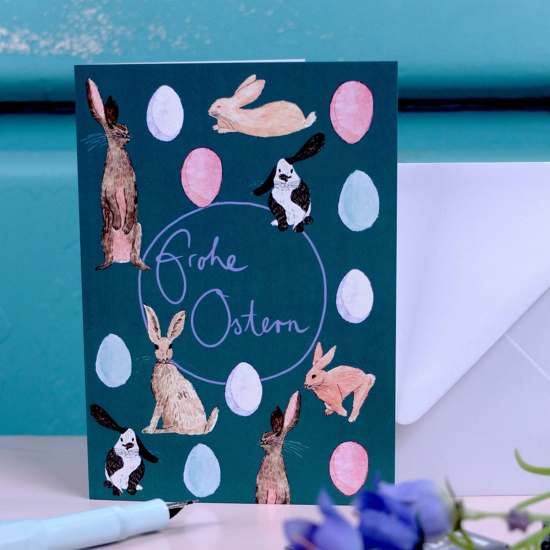 Eine Grußkarte mit illustrierten Kaninchen und pastellfarbenen Eiern auf einem blaugrünen Hintergrund. Der handgeschriebene Text in der Mitte lautet "Frohe Ostern". Ein Umschlag und violette Blumen sind in der Nähe teilweise sichtbar.