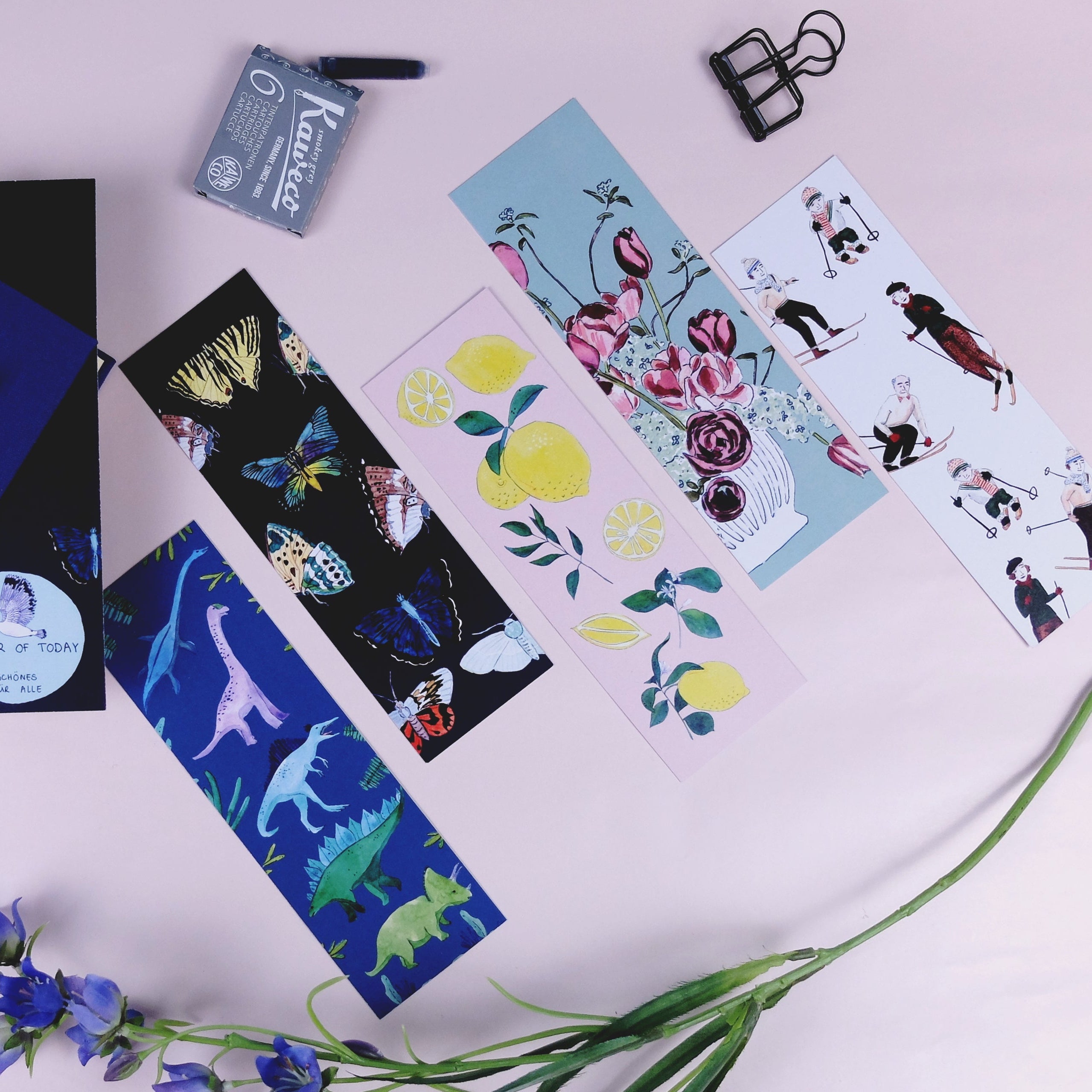 Ein blaues Notizbuch mit einem Stift, sechs farbig illustrierte Lesezeichen, ein kleiner blauer Bleistiftspitzer, eine Büroklammer und eine lila Blume sind auf einer hellrosa Fläche angeordnet.