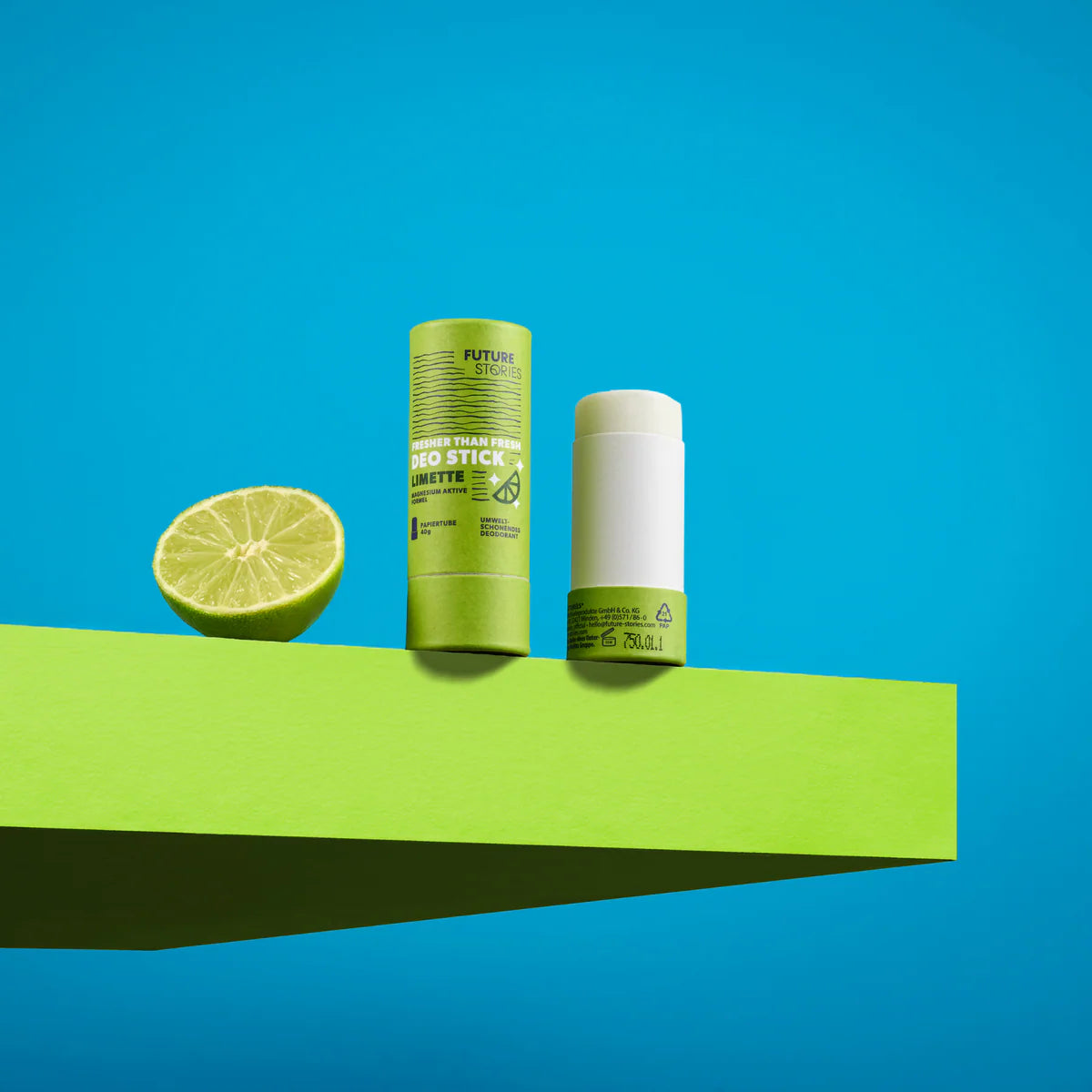 Ein grüner Deostick, dessen Kappe entfernt und daneben gelegt wurde, steht neben einer halbierten Limette auf einem leuchtend grünen Podest vor einem strahlend blauen Hintergrund.
