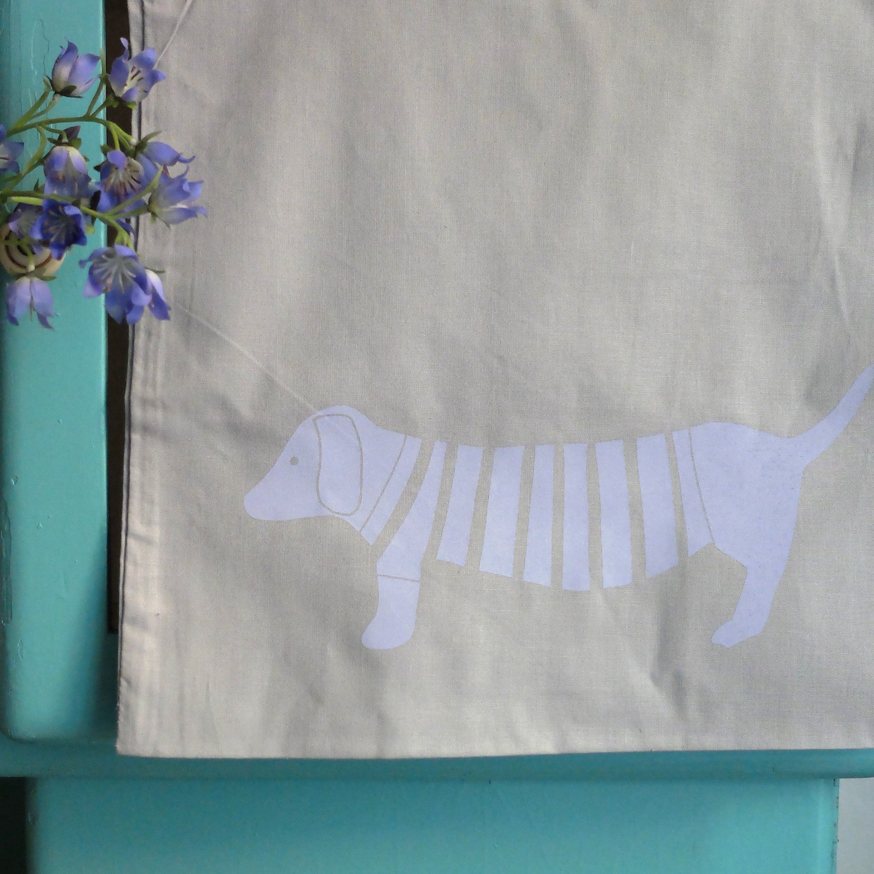 Ein weißes Tuch mit einer minimalistischen Illustration eines Dackels in einem gestreiften Pullover, daneben eine Vase mit lila Blumen, vor einem hellen, blaugrünen Hintergrund.