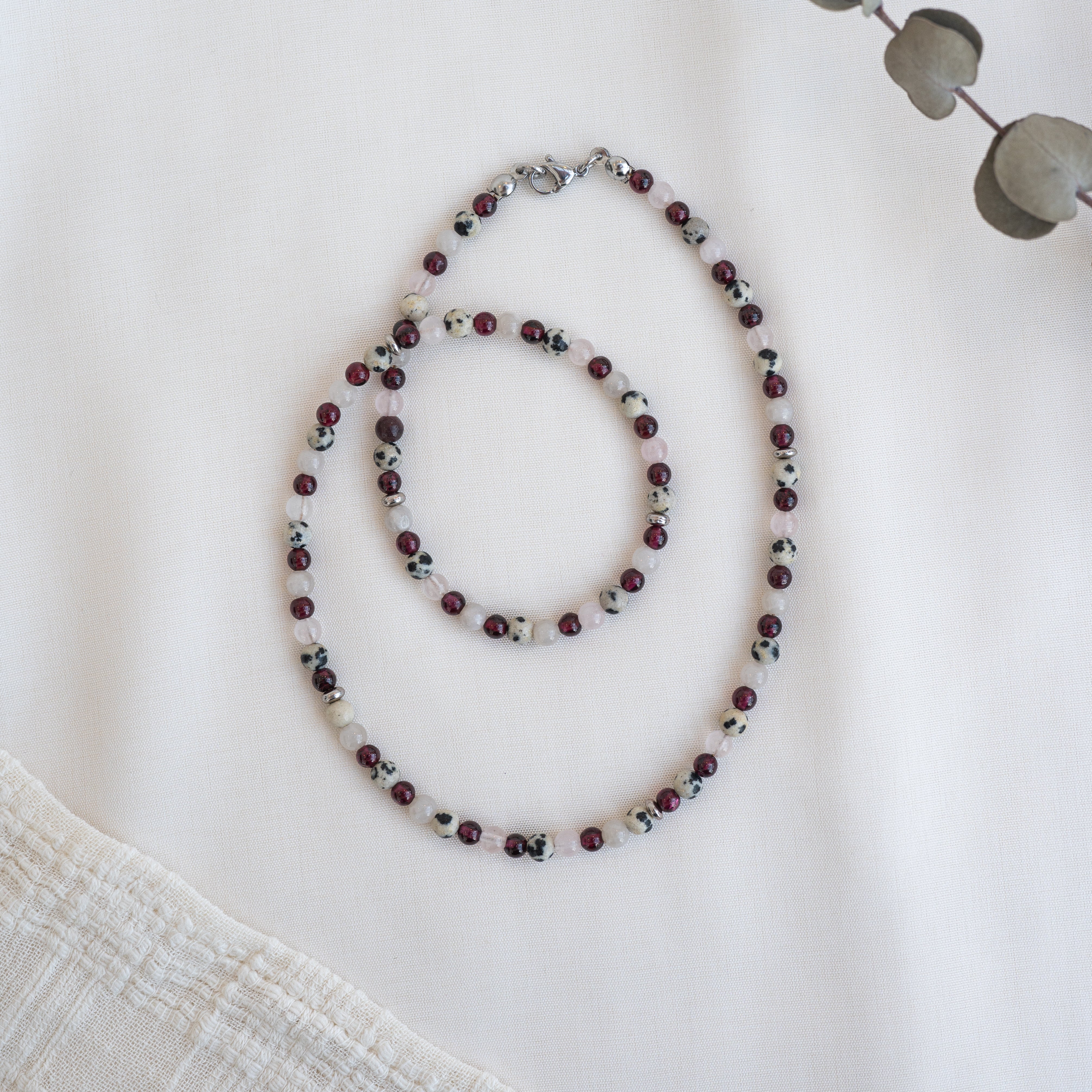 Eine Perlenkette und ein Armband mit abwechselnd roten, weißen und schwarzen Perlen sind in Kreisen auf einem weißen Stoffhintergrund angeordnet. In den Ecken sind ein beigefarbener strukturierter Stoff und getrocknete Eukalyptusblätter zu sehen.