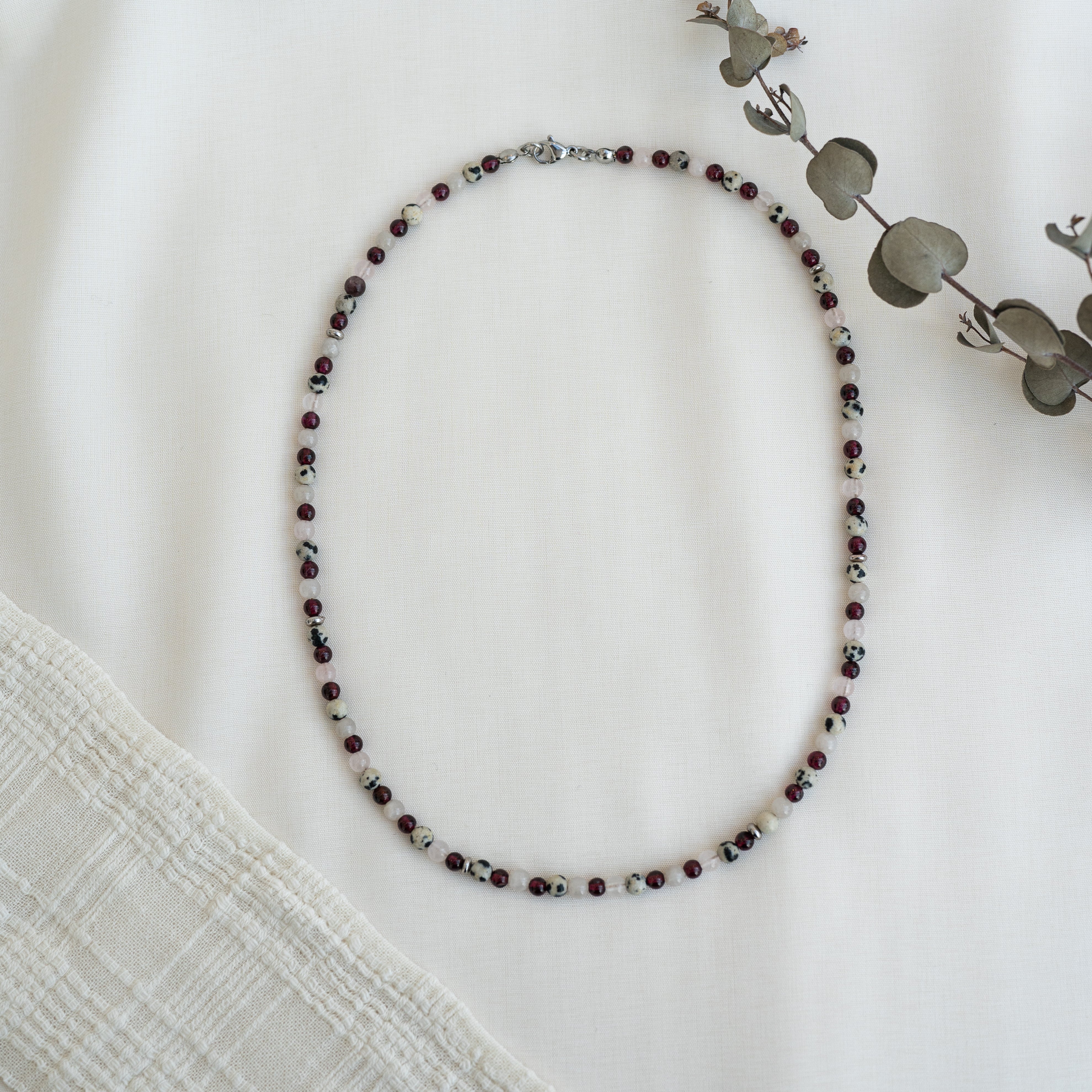 Eine Perlenkette mit abwechselnd dunkelroten und weißen Perlen liegt in ovaler Form auf einer weißen Stofffläche neben einem strukturierten Tuch und einem Zweig aus getrocknetem Eukalyptus.