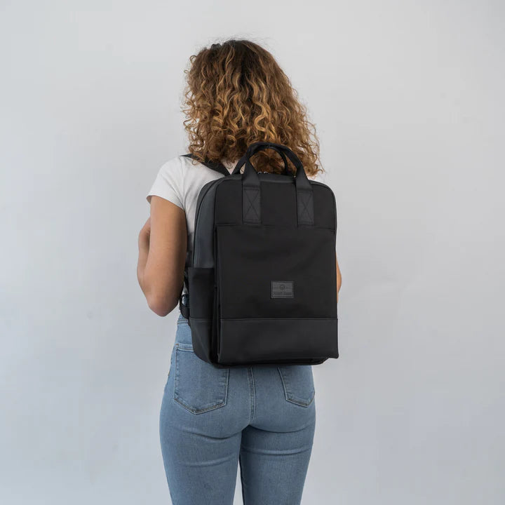 Eine Person mit lockigem Haar, die ein weißes T-Shirt und hellblaue Jeans trägt, steht von der Kamera abgewandt und modelliert einen schwarzen, quadratischen Rucksack vor einem schlichten, hellen Hintergrund.