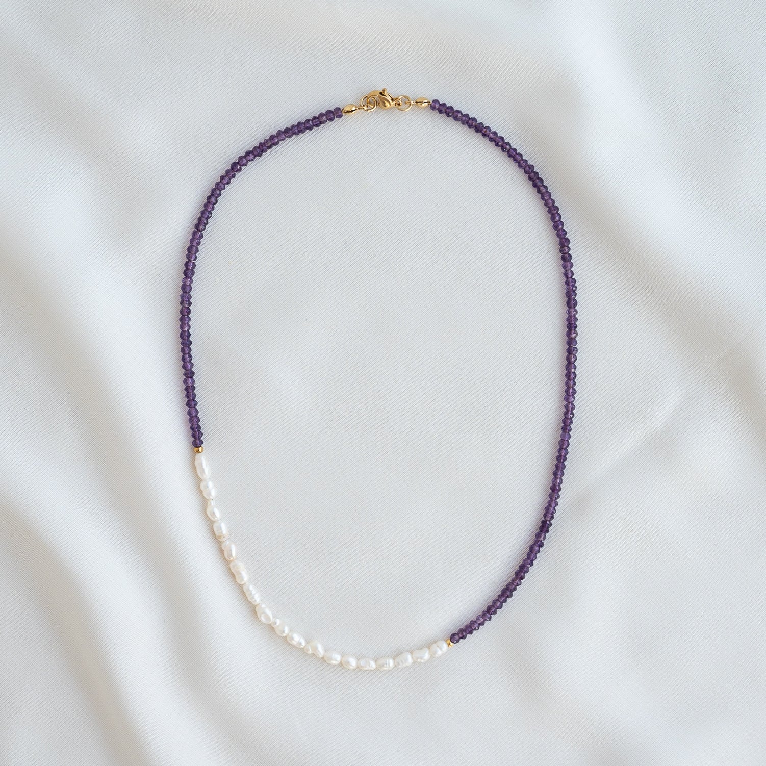 Eine zarte Halskette mit kleinen violetten Perlen auf zwei Dritteln der Länge und weißen Perlen auf dem restlichen Drittel, flach auf einem weißen Stoffuntergrund gelegt.