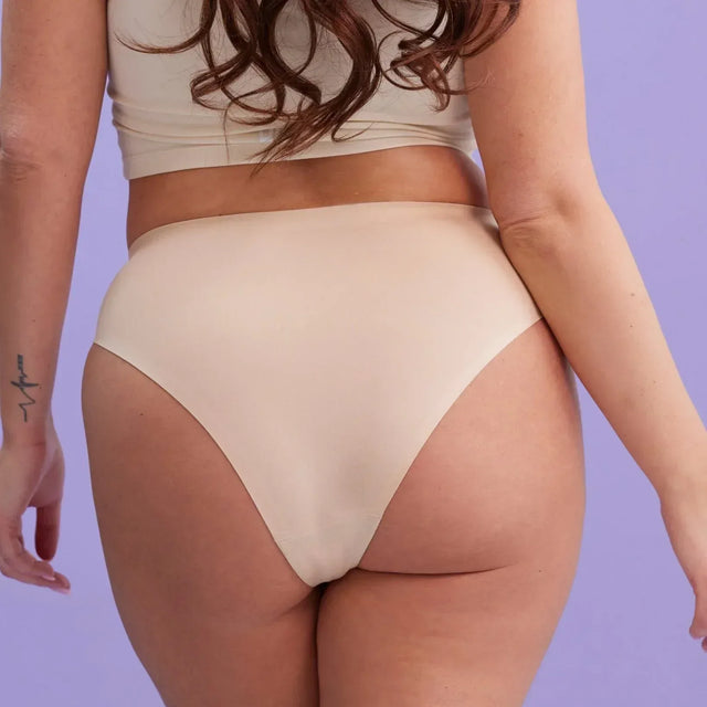 Eine Frau mit langen braunen Haaren trägt eine beigefarbene Unterwäsche mit hoher Taille und ein dazu passendes Oberteil und steht mit dem Rücken zur Kamera vor einem hellvioletten Hintergrund. Auf ihrem linken Arm ist eine kleine Tätowierung zu sehen.